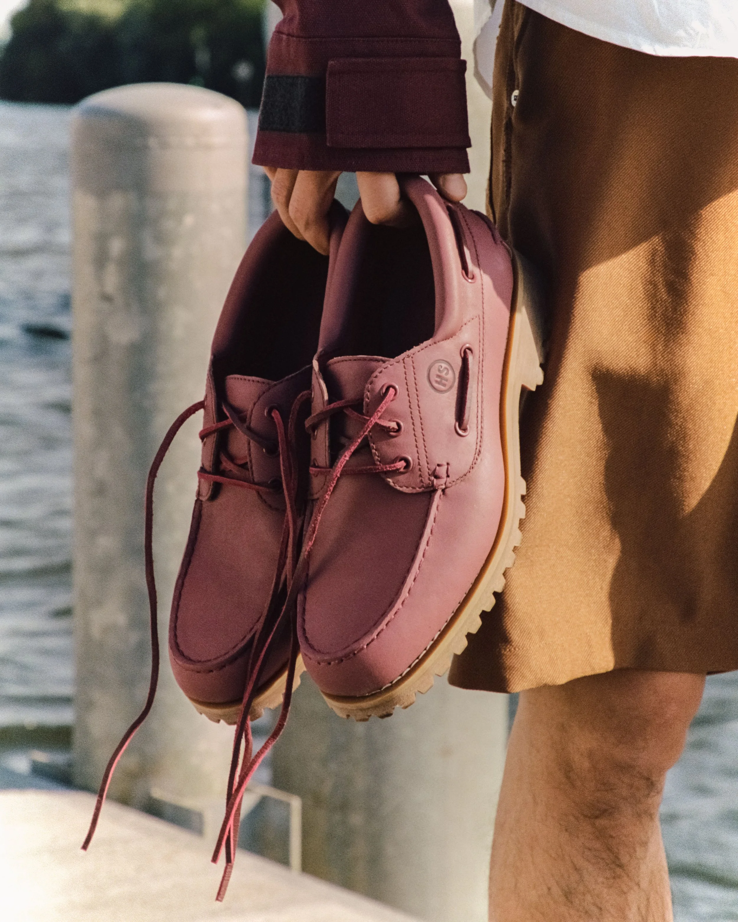 A person holding a pair of pink boat shoes outdoors near water, with a wooden dock and a metal post visible in the background.