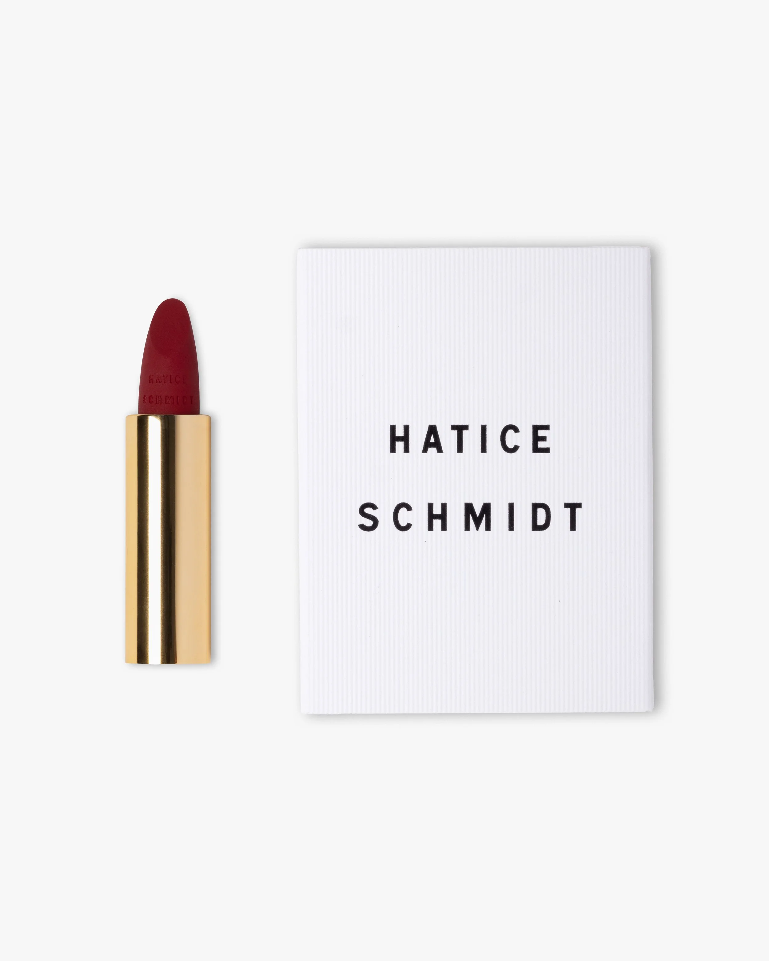 Red lipstick in a gold tube and a white box with black text reading 'HATICE SCHMIDT' on a white background.