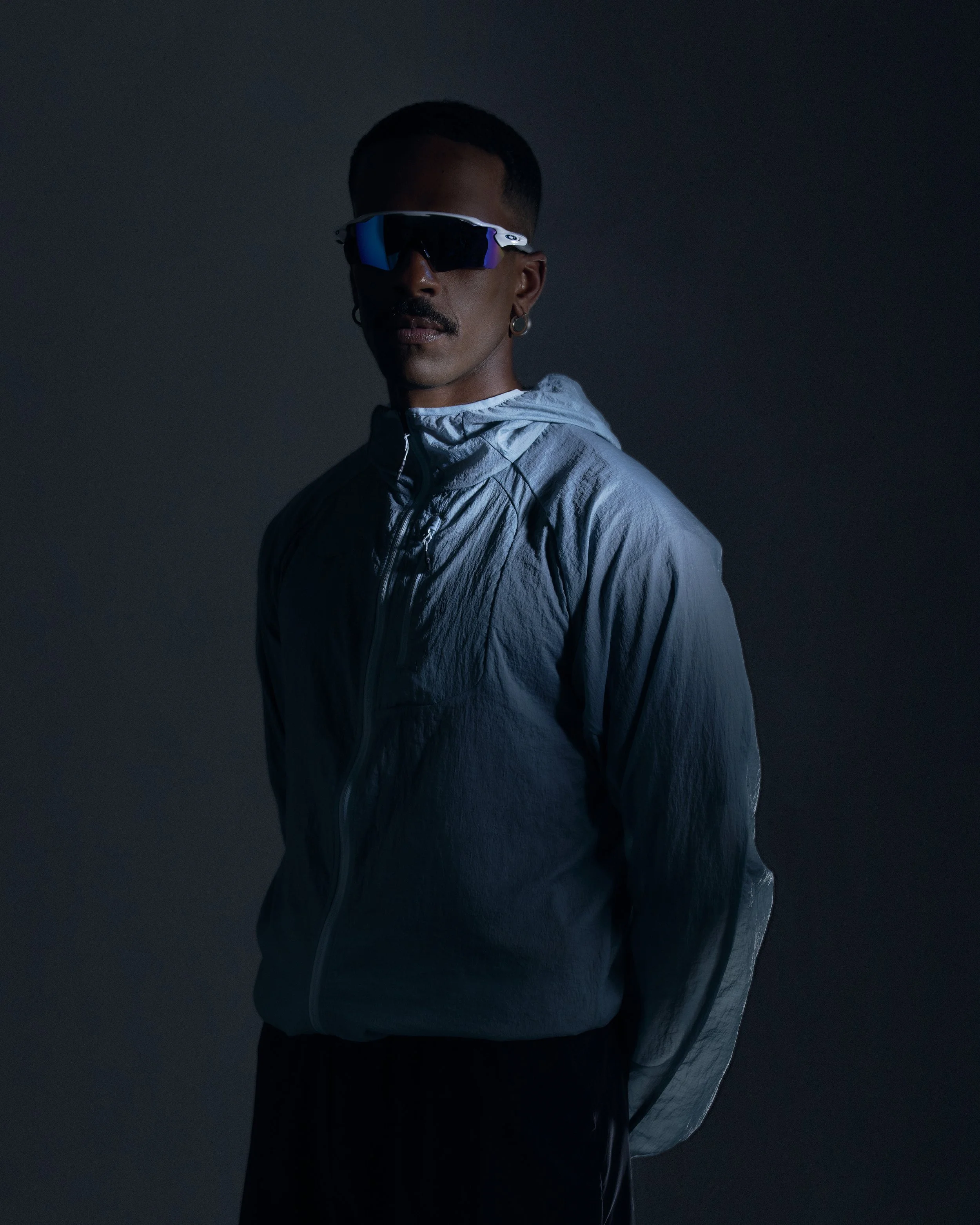 A man wearing sunglasses and a light blue windbreaker jacket, standing against a dark background.
