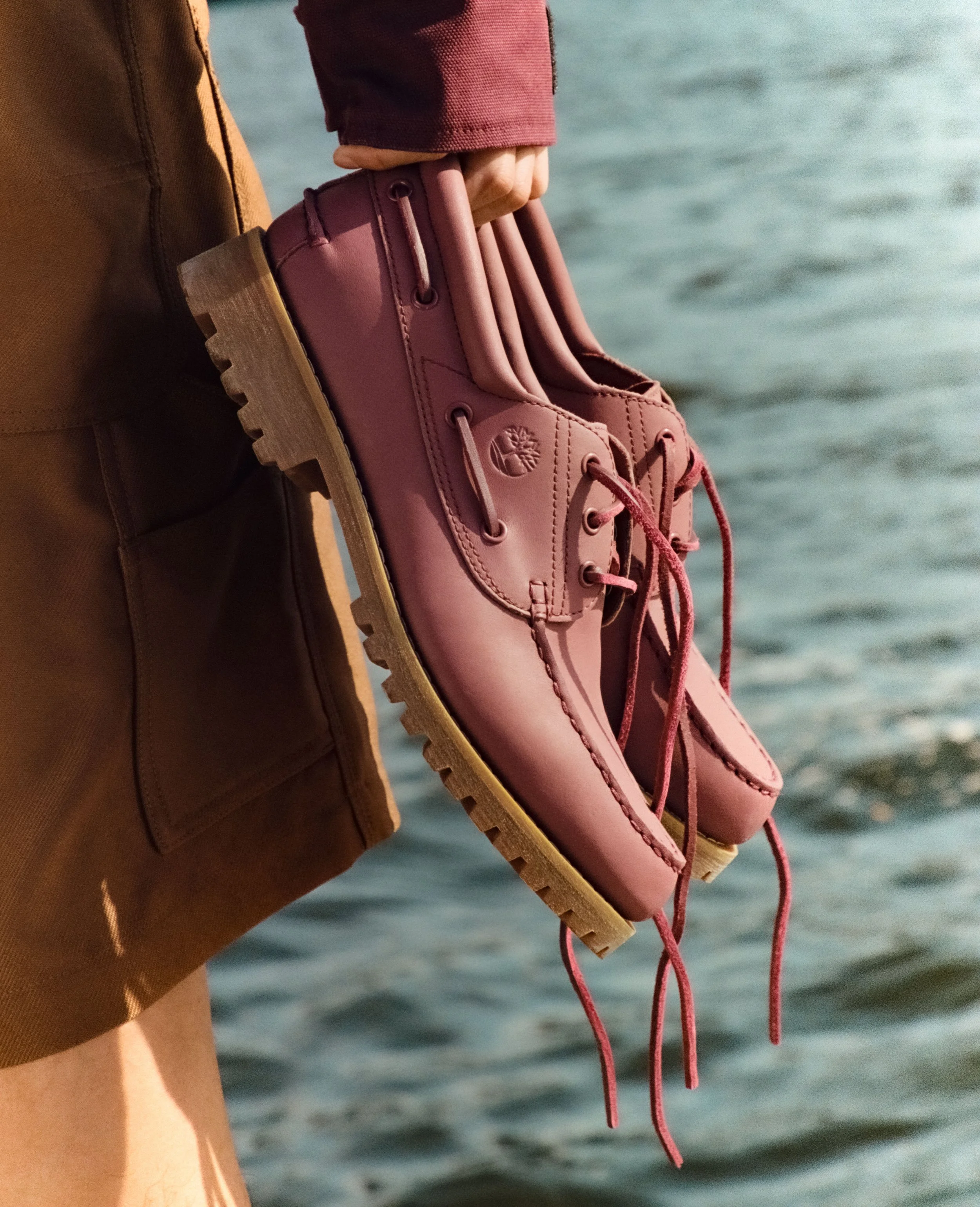 Close-up of a person's hand holding pink Timberland-style boots over a body of water, with a brown jacket partially visible.