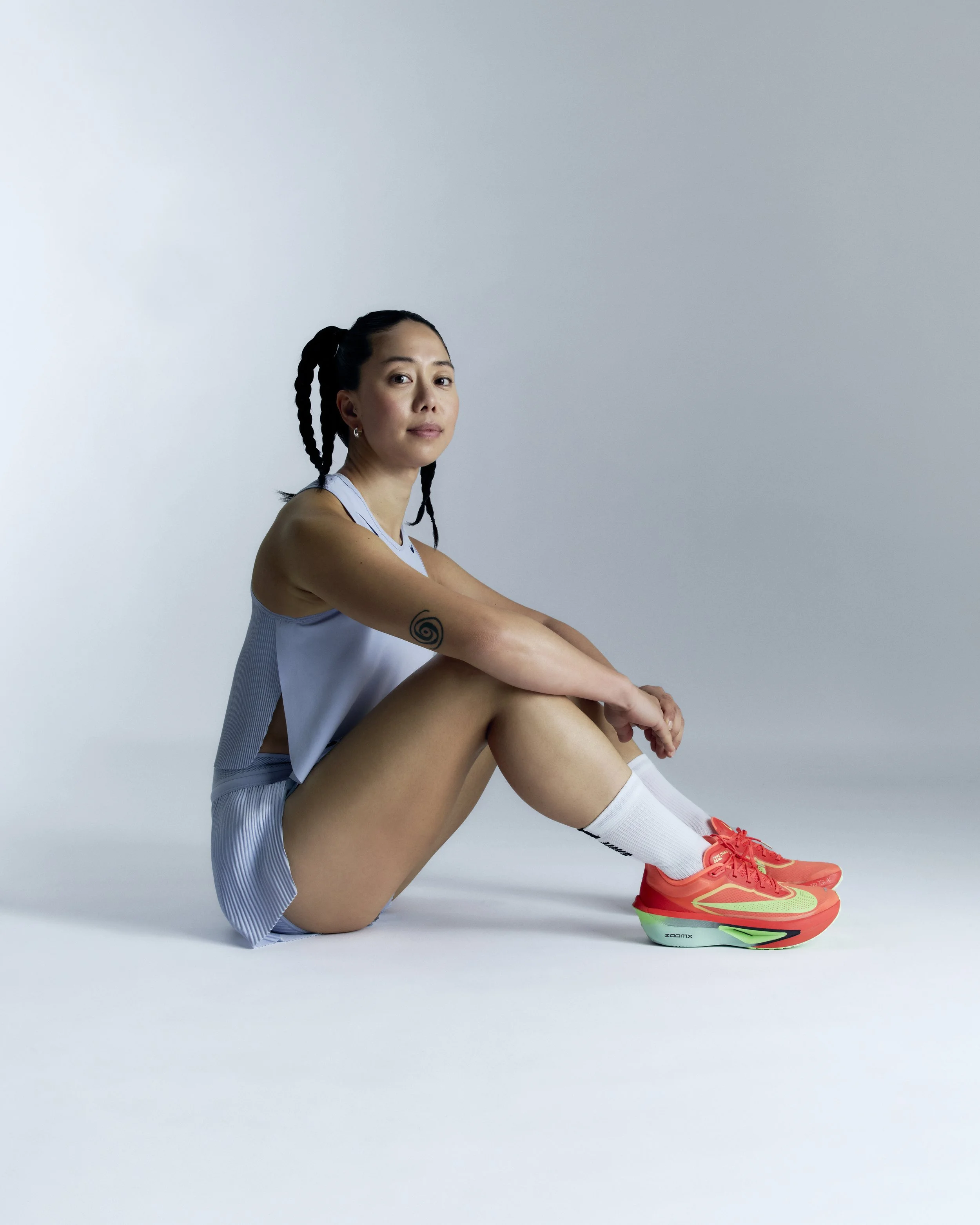A young woman sits on the floor against a plain, light-colored background, wearing athletic clothing and bright coral sneakers, with her arms resting on her knees and looking at the camera.