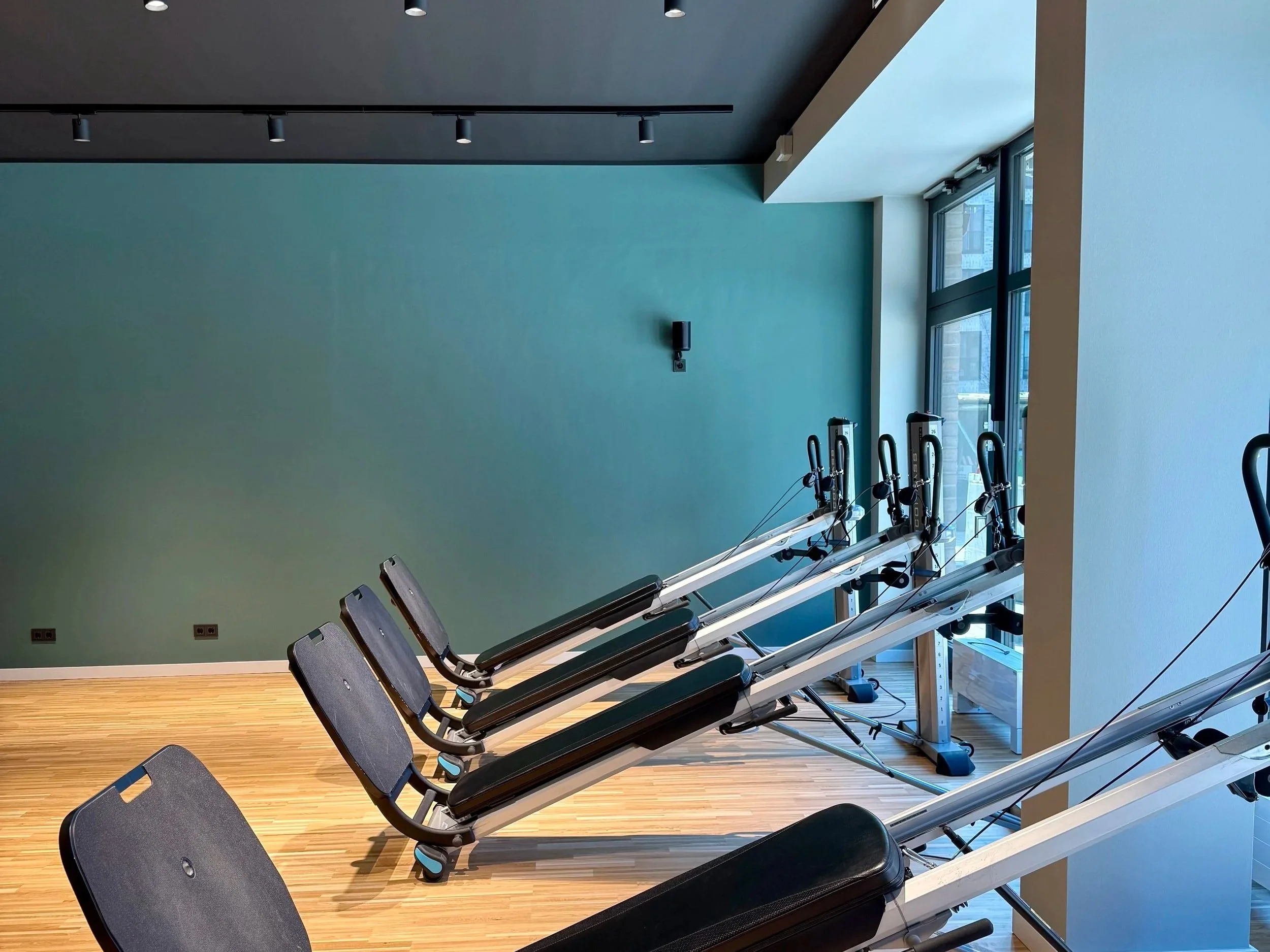 Innenraum eines Fitnessstudios mit mehreren Total Gym Trainingsgeräten auf Holzfußboden, große Fenster, blau-grüne Wand, schwarze Decke, Deckenleuchten und einem Wandlautsprecher.