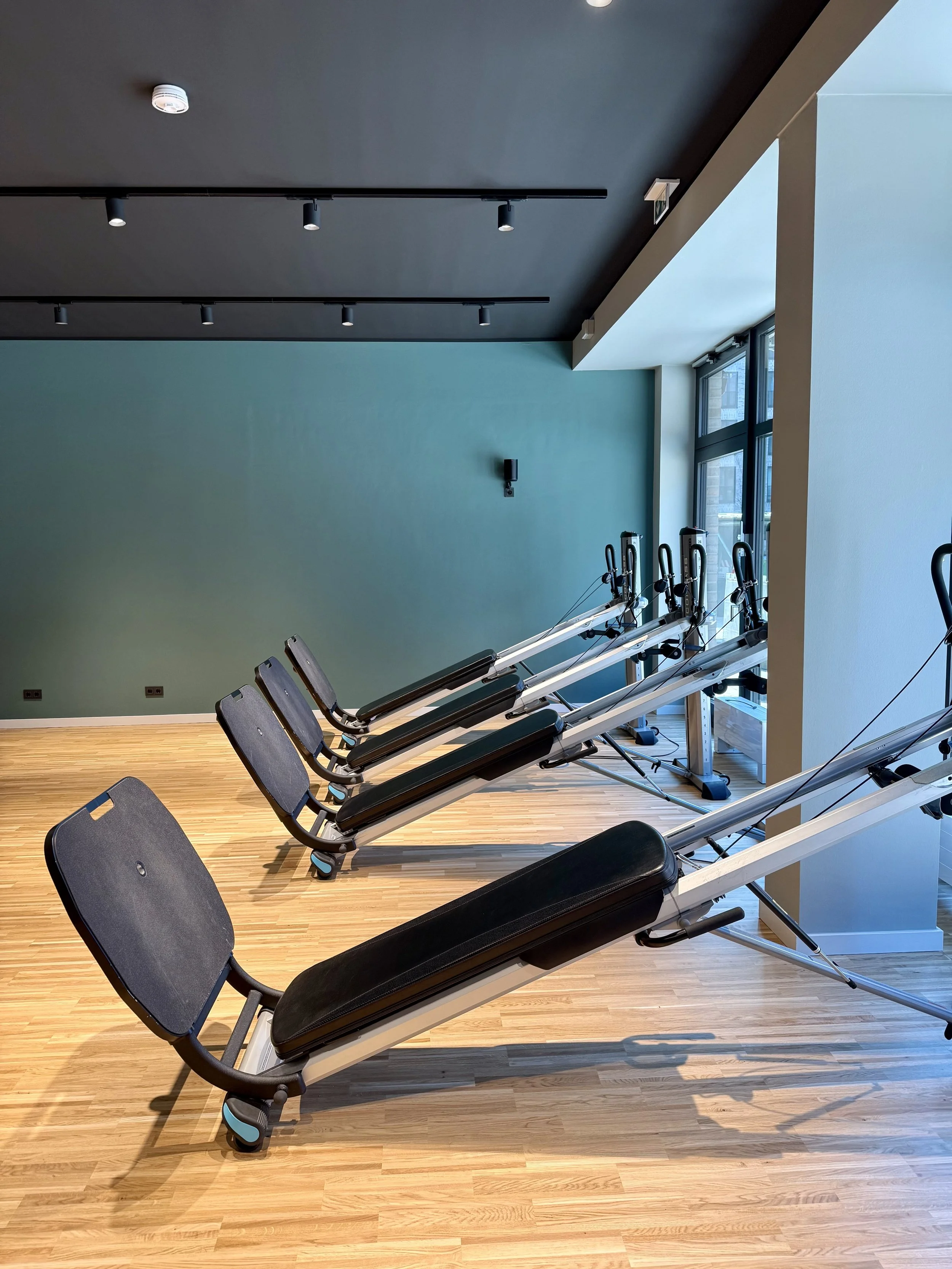 Vier Liege- und Trainingsgeräte in einem Fitnessraum mit Holzfußboden und großem Fenster.