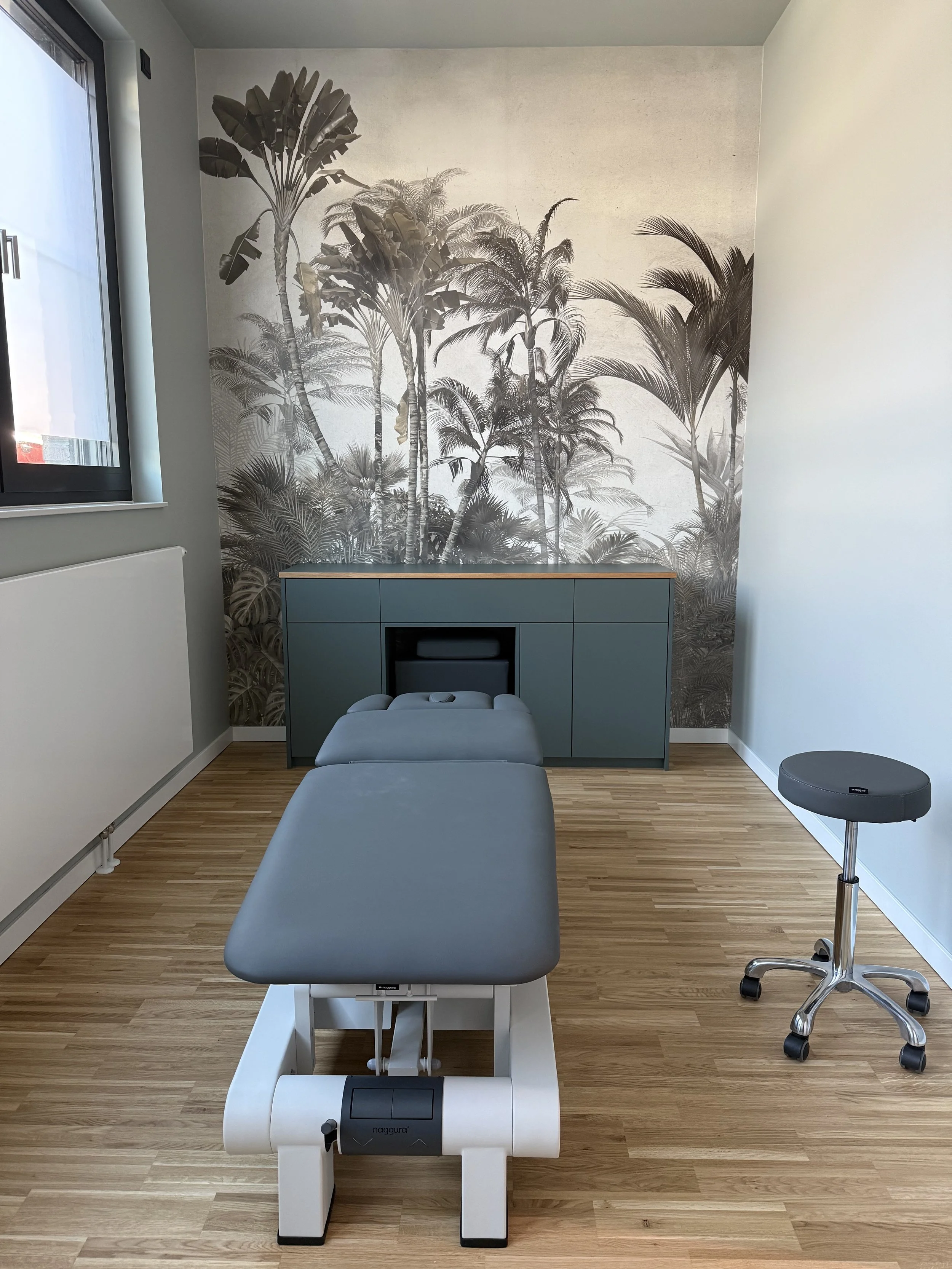Ein Behandlungszimmer mit einer Massageliege, einem Stuhl auf Rollen, einem Schrank vor einer Wand mit Palmen- und Baumtapete, Sonnenlicht durch ein Fenster auf der linken Seite.