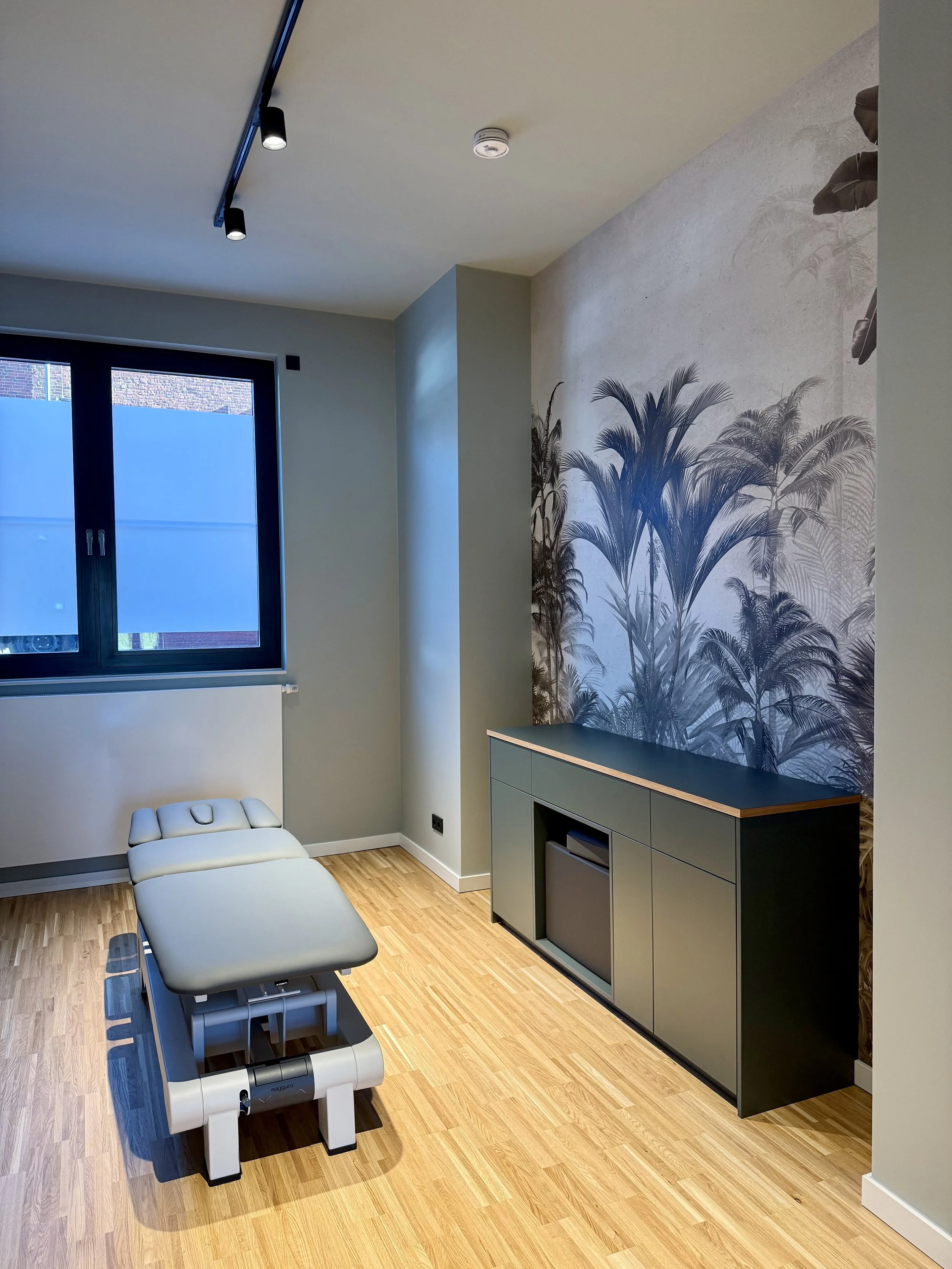 Ein Behandlungszimmer mit Untersuchungsliege, einem Fenster, einer Wand mit einer Palme-Dekor, und einem schwarzen Schrank.