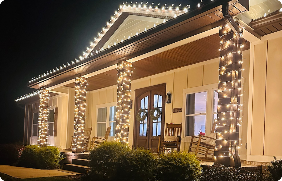 A house decorated with string lights on the porch, with rocking chairs and wreaths on the door, illuminated at night.