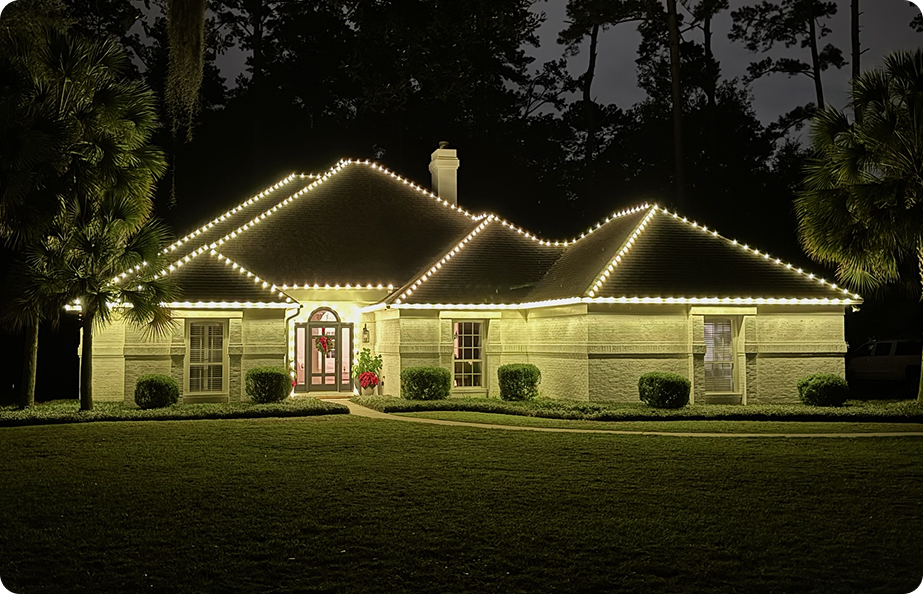 A house decorated with string lights outlining its roof at night, surrounded by a lawn and trees.