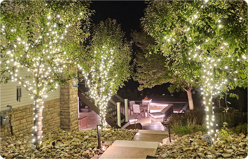 Nighttime outdoor patio decorated with trees wrapped in string lights, with seating area and hot tub in the background.