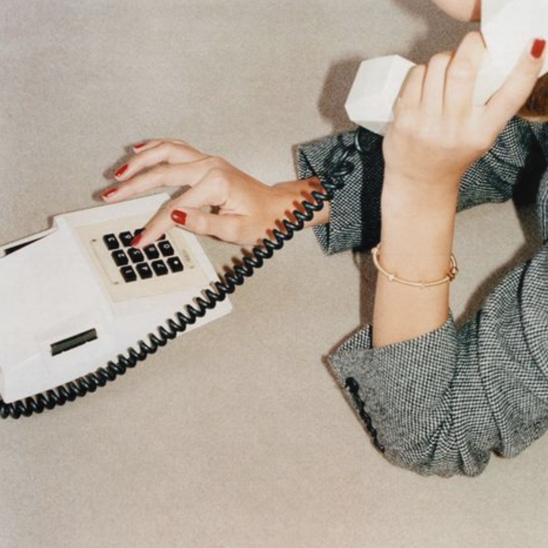 Person with red-painted nails holding a landline telephone receiver, with other hand on the phone's dial pad, wearing a checkered blazer and a gold bracelet.