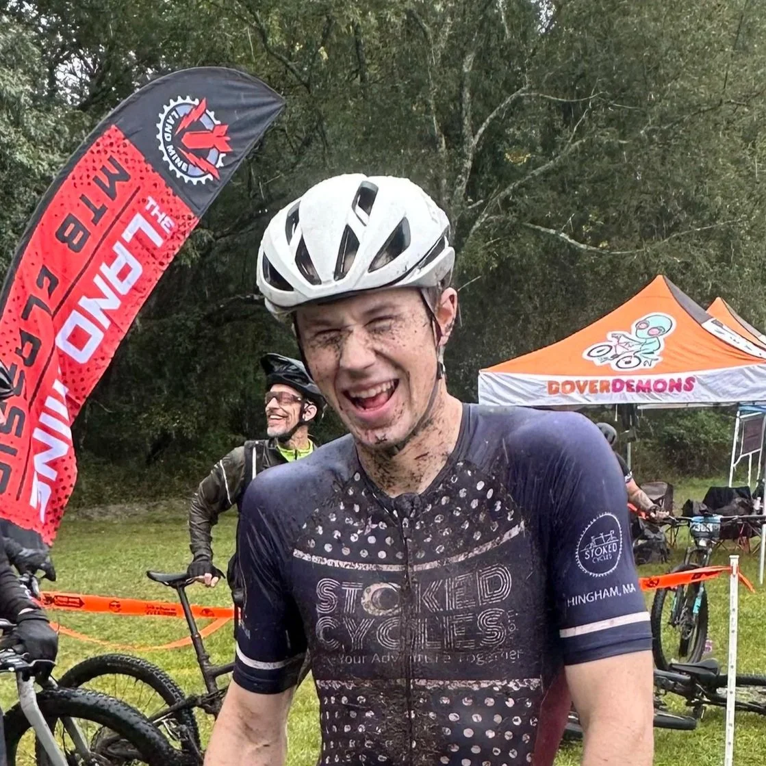 A smiling male cyclist with a muddy face, wearing a white helmet and a black cycling jersey, standing outdoors near a red and black flag and an orange tent. Other cyclists and bikes are visible in the background.