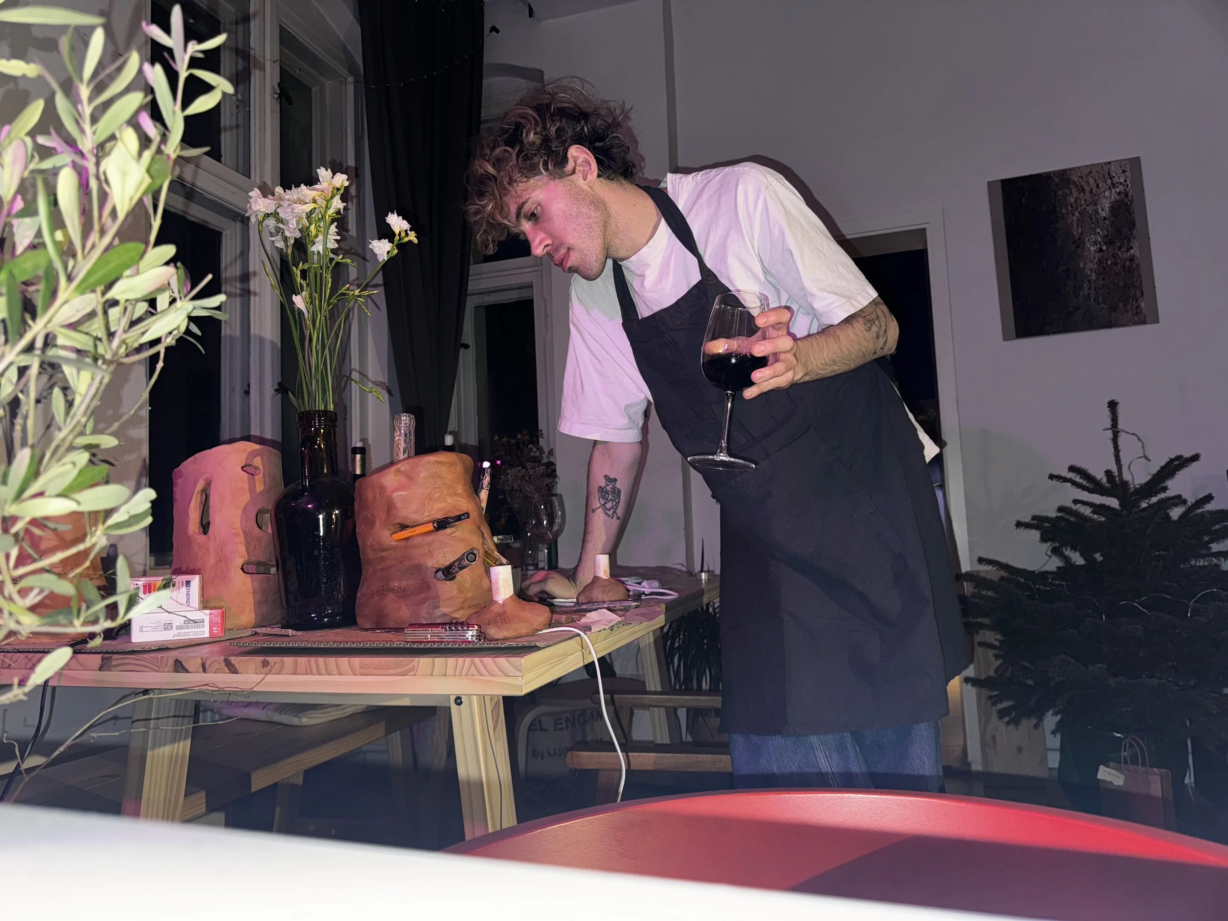 A young man with curly hair wearing a white t-shirt and black apron holding a glass of red wine while leaning over a table with pottery, bottles, and tools. There is a window with dark curtains, some artwork on the wall, and a Christmas tree in the background.