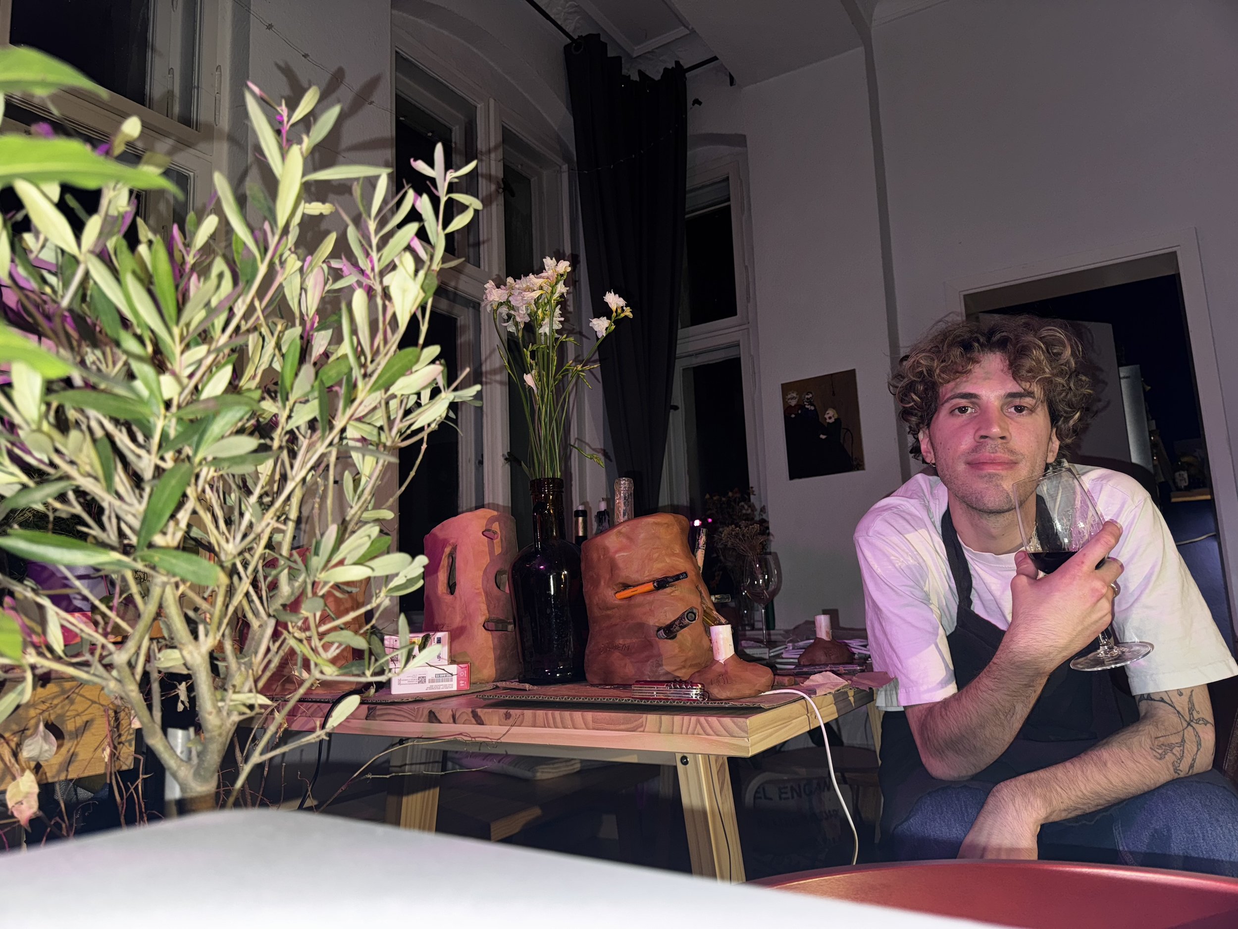 Seated at a workbench, shaping a clay lamp by hand, surrounded by tools, sketches, and unfinished pieces in a studio filled with natural light.