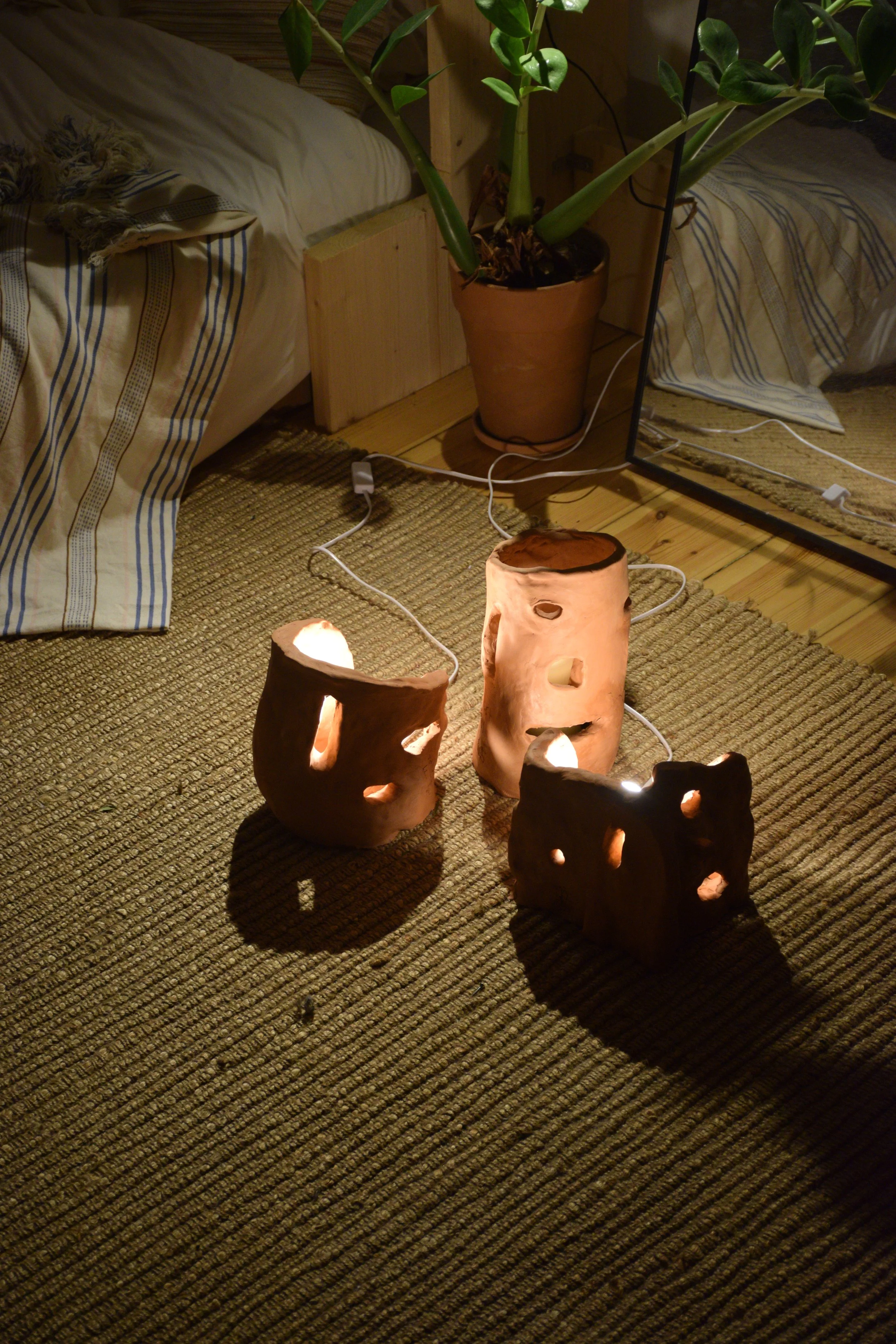 Decorative candle holders made from hollowed-out logs with carved faces, illuminated from within, placed on a textured carpet in a cozy room with a bed, mirror, potted plant, and electrical cords.