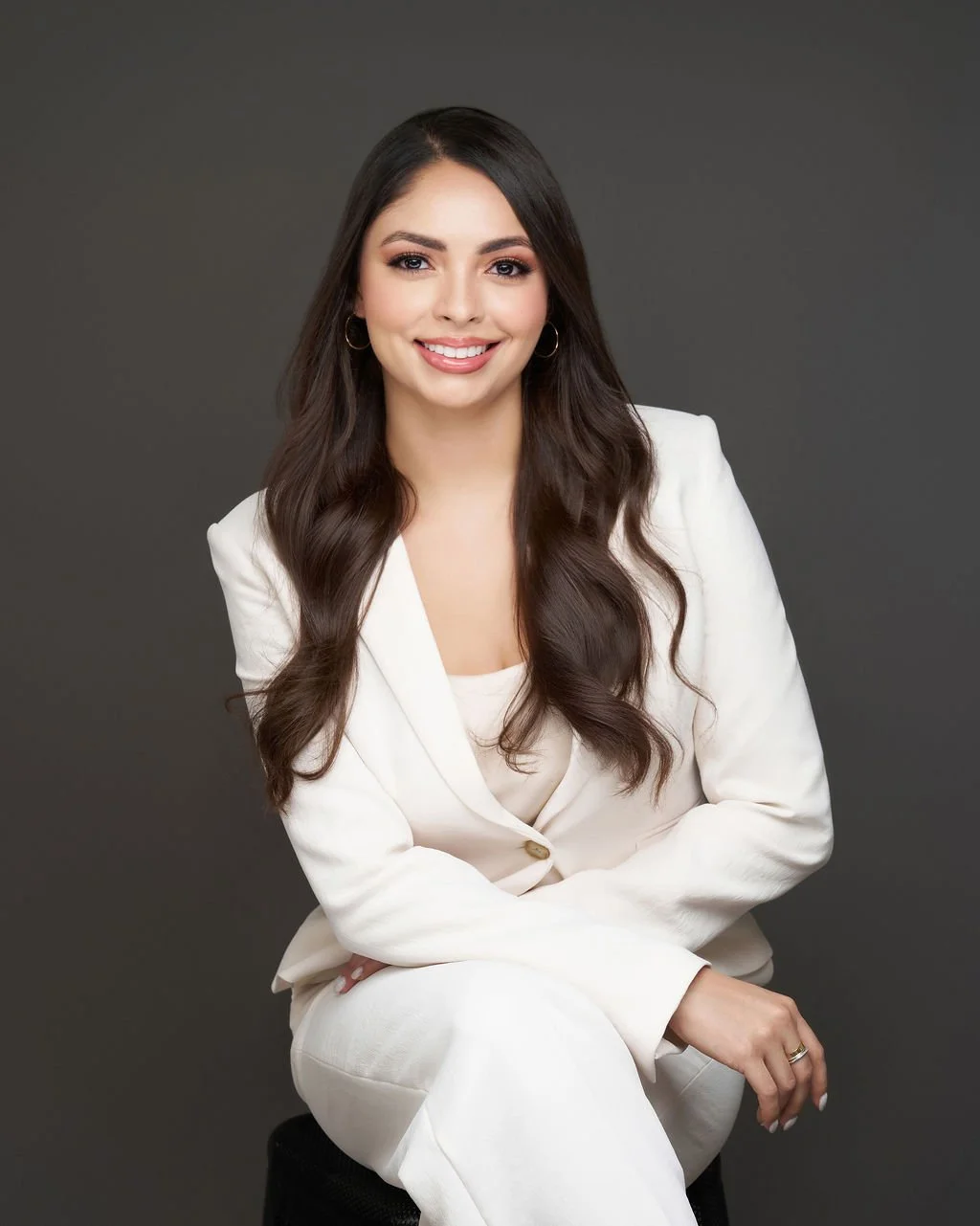 Portrait of a young woman with long dark hair, wearing a white suit, smiling, against a dark gray background.