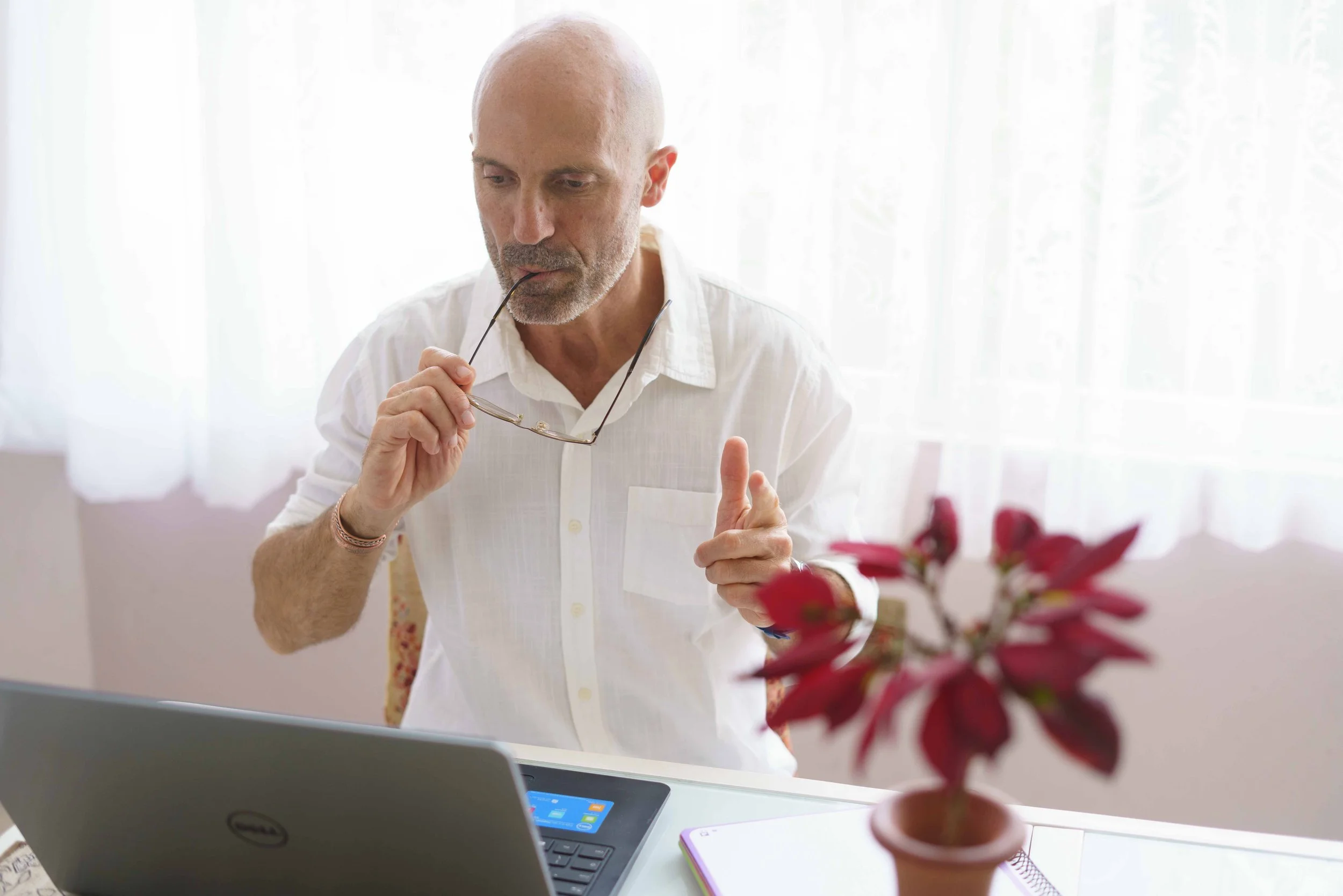 Hombre mayor sentado en una mesa de oficina con una laptop, sosteniendo unas gafas y tocándose la vista, en un ambiente con cortinas blancas y planta decorativa en primer plano.
