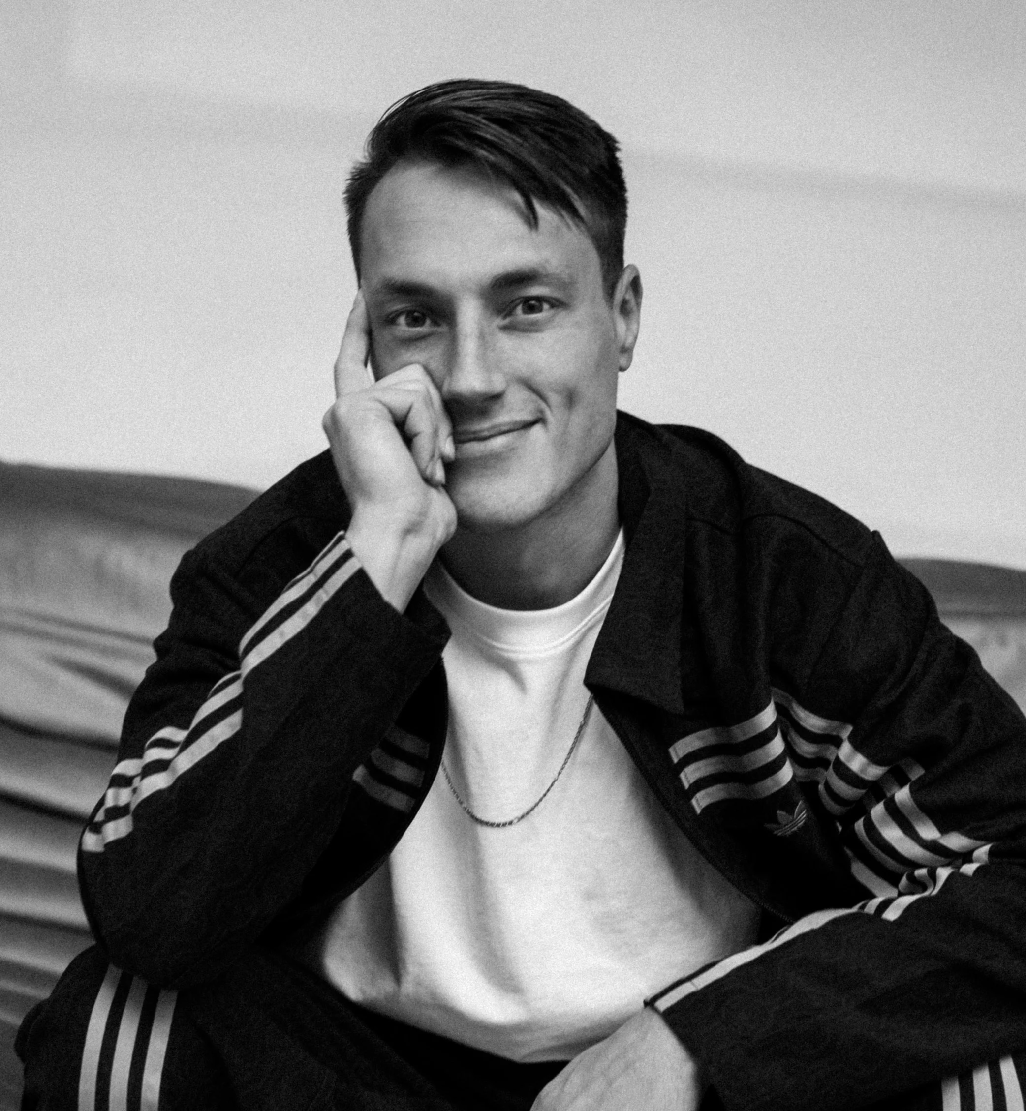 A young man with short dark hair sitting on a bench, resting his chin on his hand, smiling at the camera.