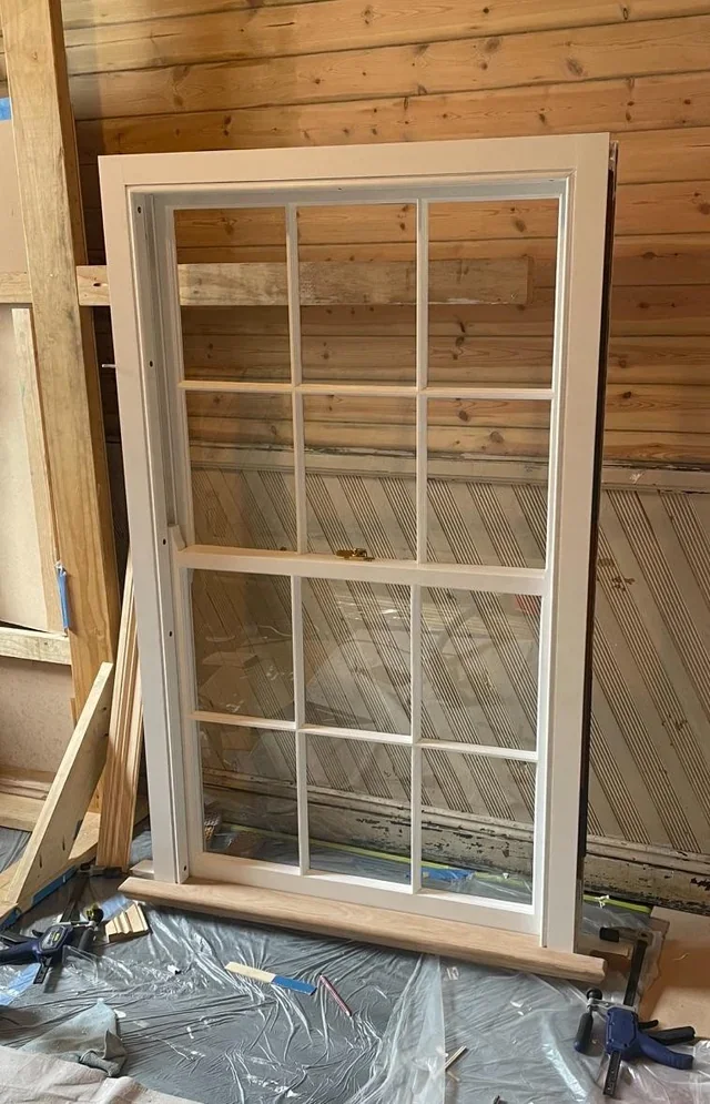 Newly built white-framed window with multiple panes in a room under construction, with wooden walls and construction tools on a plastic-covered floor.