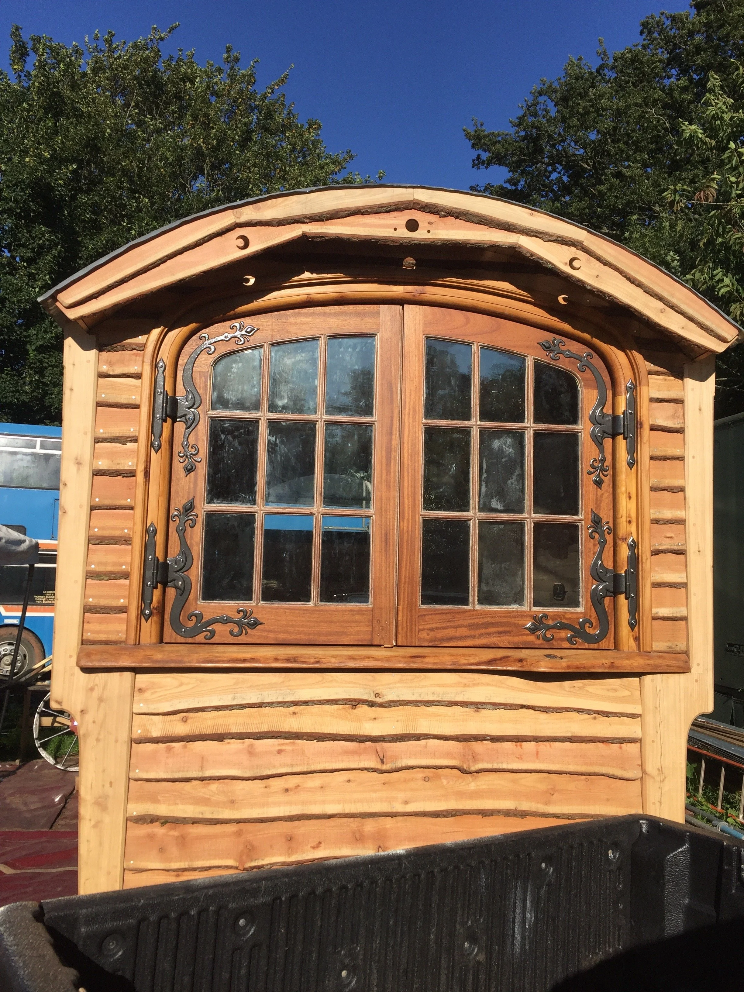 A shepherds hut with decorative black hinges and a curved roof, featuring a window with nine panes and a natural finish, situated outdoors under a bright blue sky with green trees in the background.