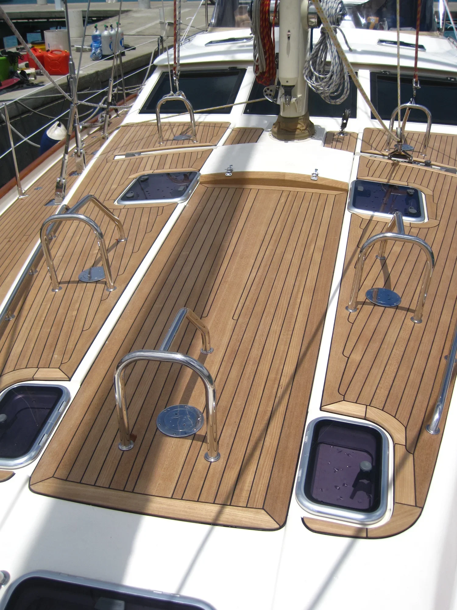 The deck of a sailboat with wooden flooring, stainless steel stanchions, and hatches, docked at a marina.