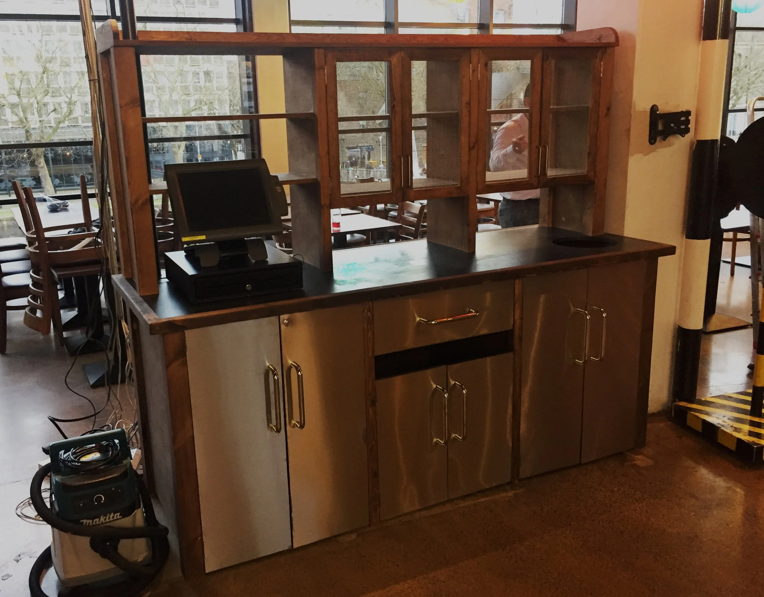A commercial kitchen or bar station with a wooden and stainless steel setup. The station has a countertop, glass-front cabinets above, a point of sale system, a sink, and a vacuum cleaner nearby. Large windows in the background let in natural light, and seating is visible outside.