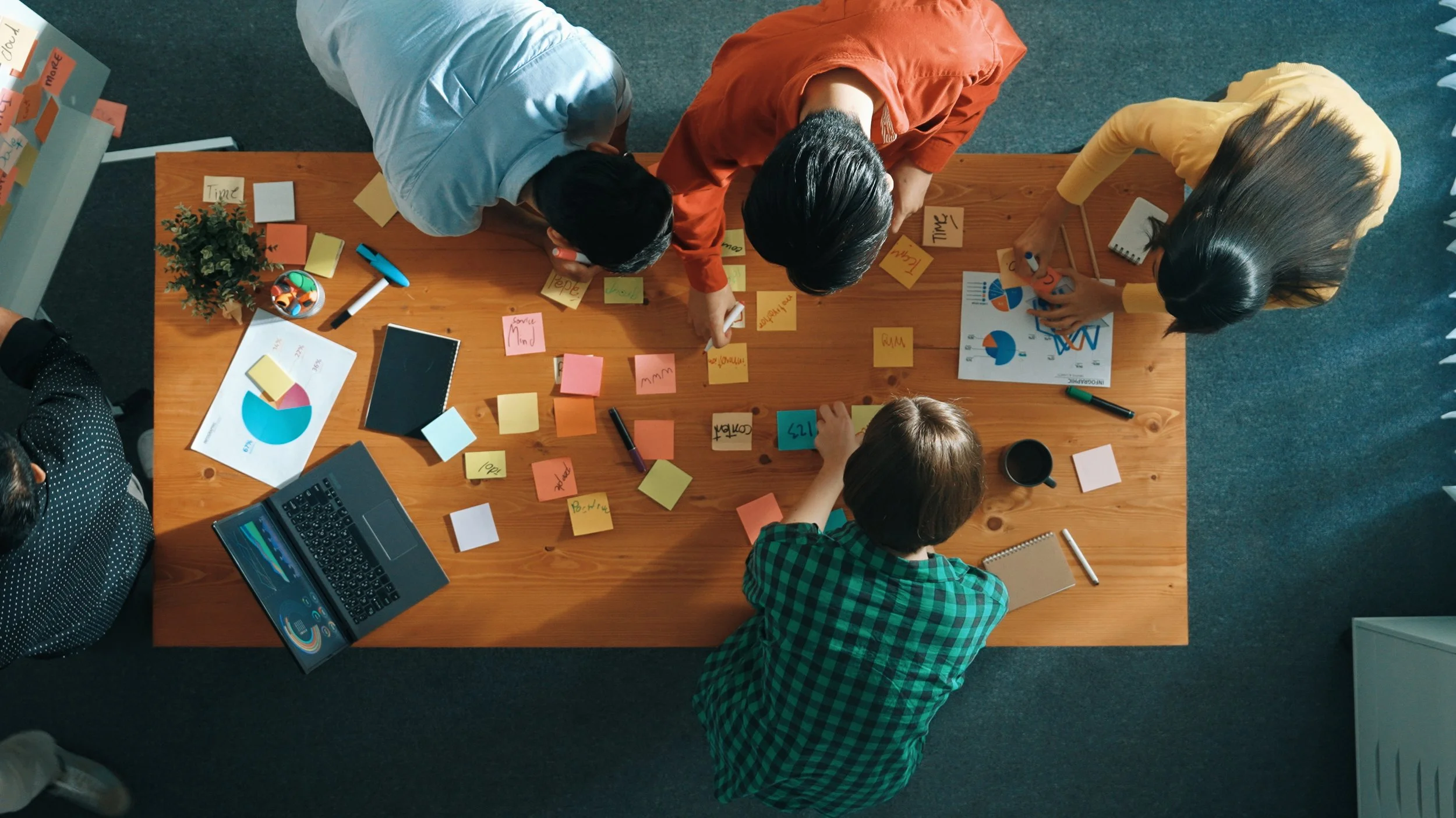 A top-down view of five people collaborating at a wooden table covered with sticky notes, documents, a laptop, and writing utensils during a meeting or brainstorming session.