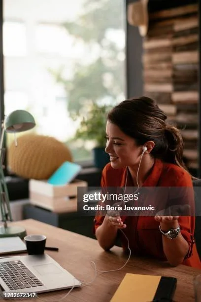 A woman smiling and talking to herself or video call while wearing earphones and sitting at a desk in a bright room with a window, green lamp, books, and a yellow pillow.