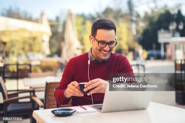 Man sitting at an outdoor cafe table, smiling while looking at his phone. He has dark hair, glasses, and is wearing a red sweater with earphones. There is a laptop and a small dish on the table.