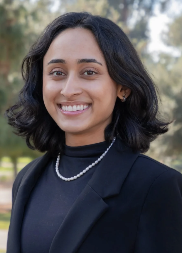A woman with shoulder-length dark hair, wearing a black blazer, pearl necklace, and earrings, smiling outdoors with blurred trees in the background.