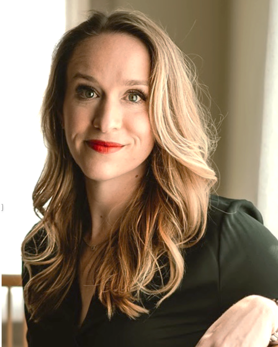 A woman with long, wavy, light brown hair, green eyes, and red lipstick, smiling slightly, dressed in a black top, sitting near a window.