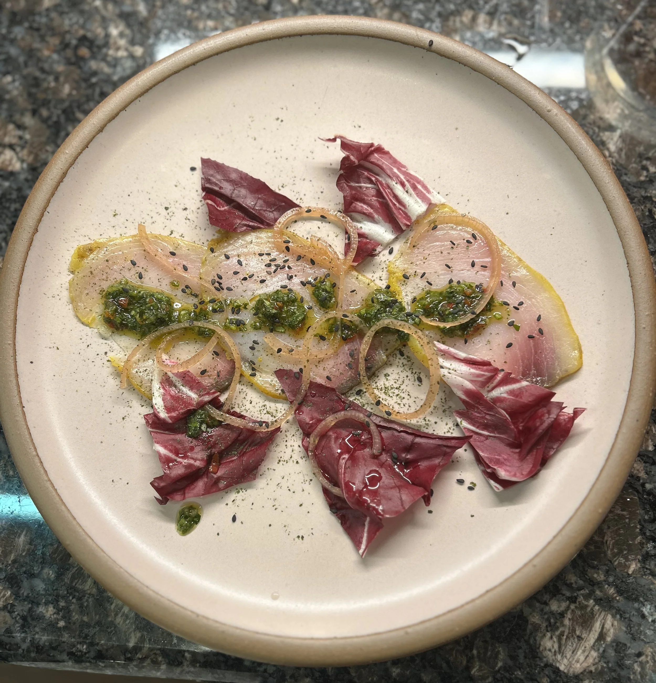 A bowl of thinly sliced raw fish garnished with seasonings, herbs, and black sesame seeds, decorated with radicchio leaves and onion slices, on a granite countertop.