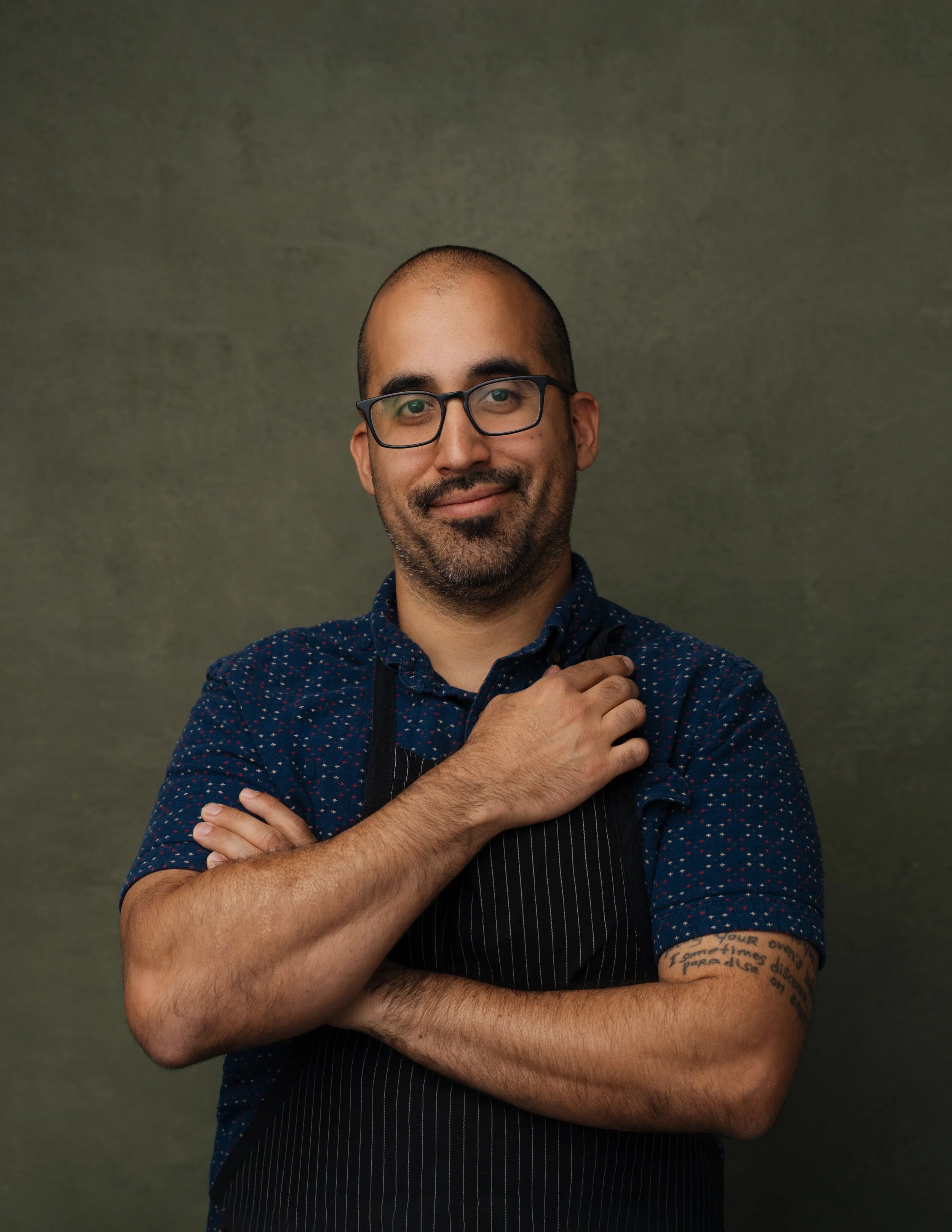 A man with glasses, a trimmed beard, and a tattoo on his left forearm standing against a plain green background. He's wearing a dark, dotted shirt with a black apron and smiling.