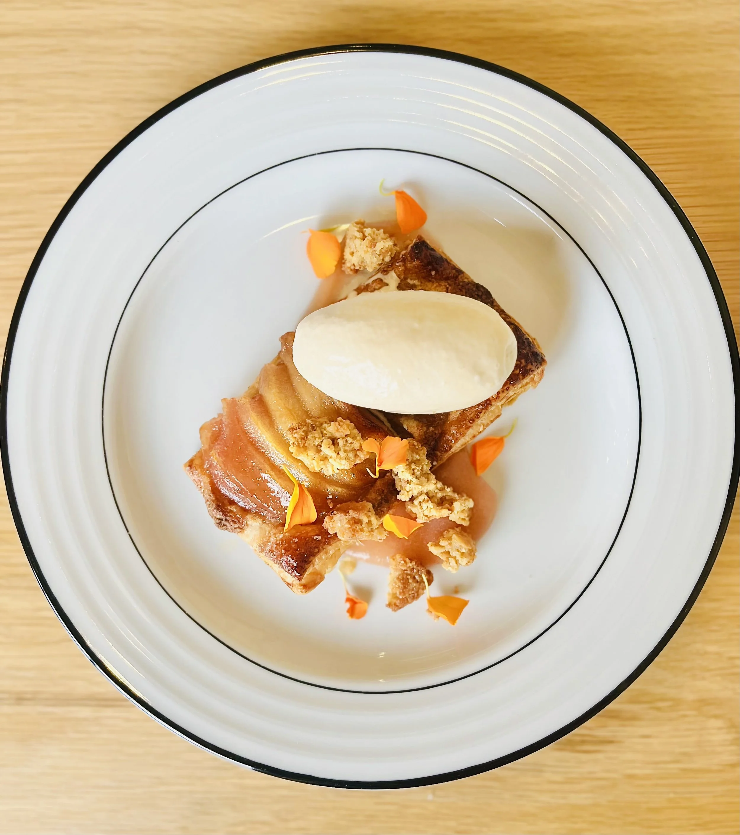 A plated dessert with a piece of caramelized apple tart, a scoop of vanilla ice cream, and edible orange flower petals, served on a white plate with black rim on a wooden surface.