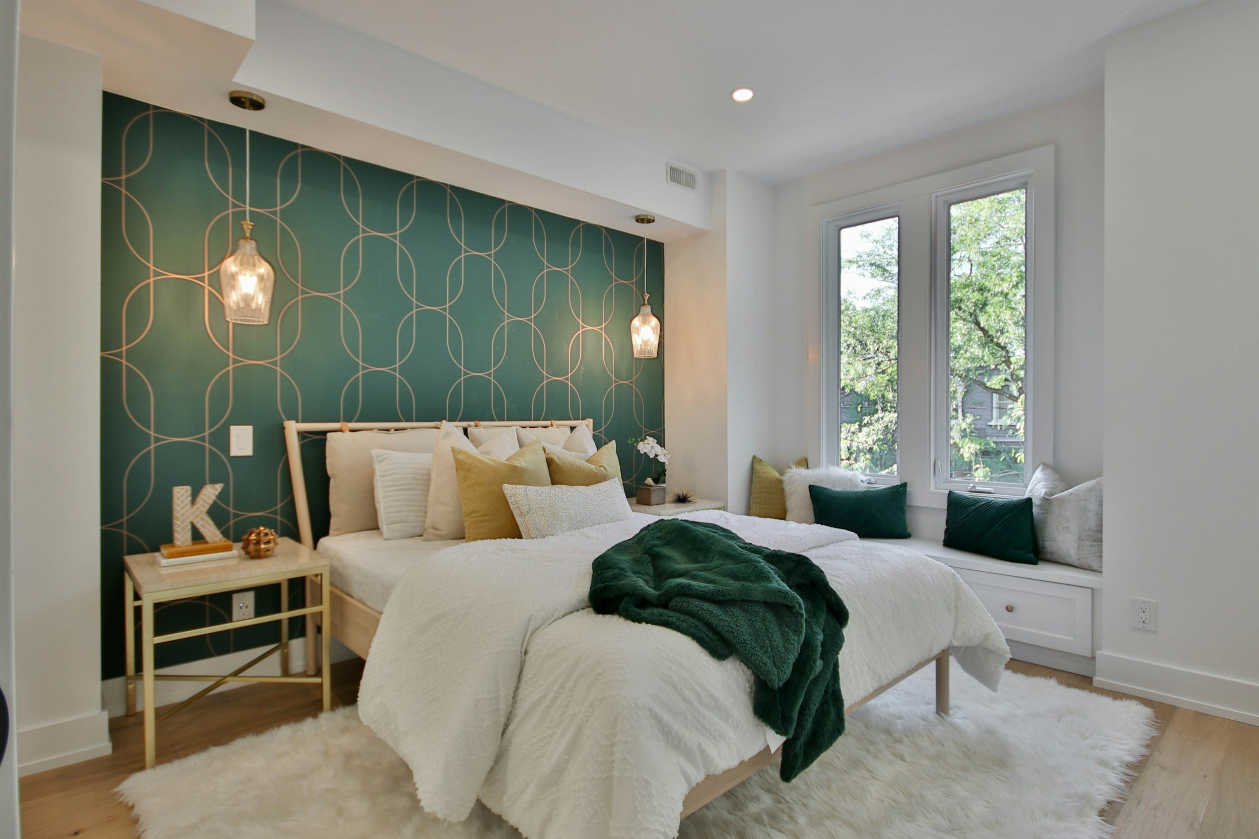 A modern bedroom with a white bed, gold and beige pillows, green throw blanket, and a blue accent wall with geometric gold pattern. There are hanging pendant lights, a gold side table with decorative items, and a window seat with white and green cush
