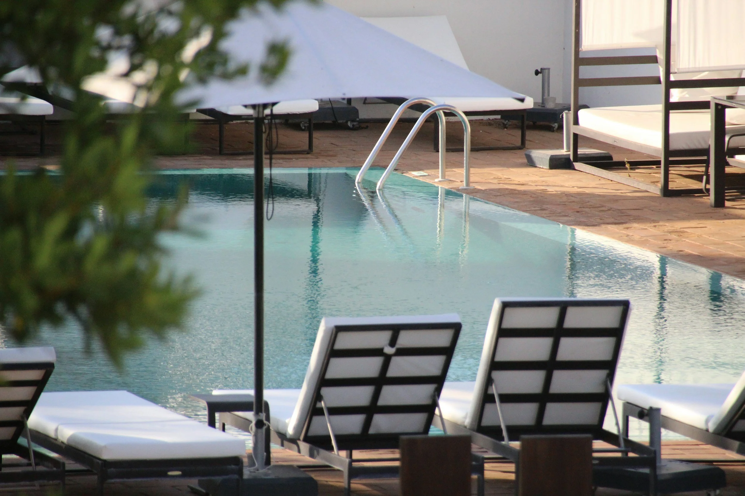 Empty swimming pool with a metal ladder, poolside lounge chairs, and a large umbrella on a brick deck.