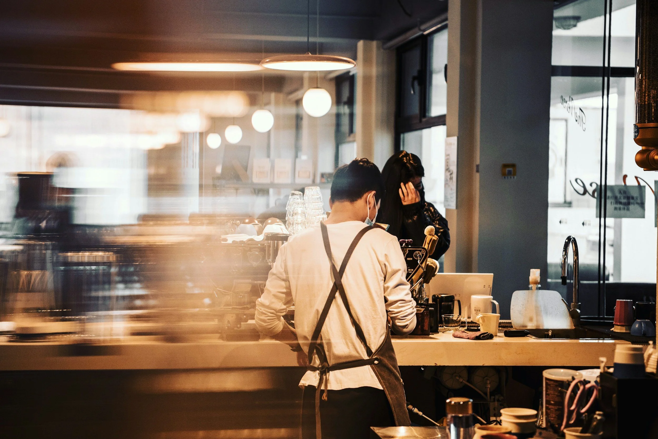 A barista preparing drinks behind a counter in a coffee shop, while a customer wearing a mask orders at the counter. The scene is warm and softly lit.