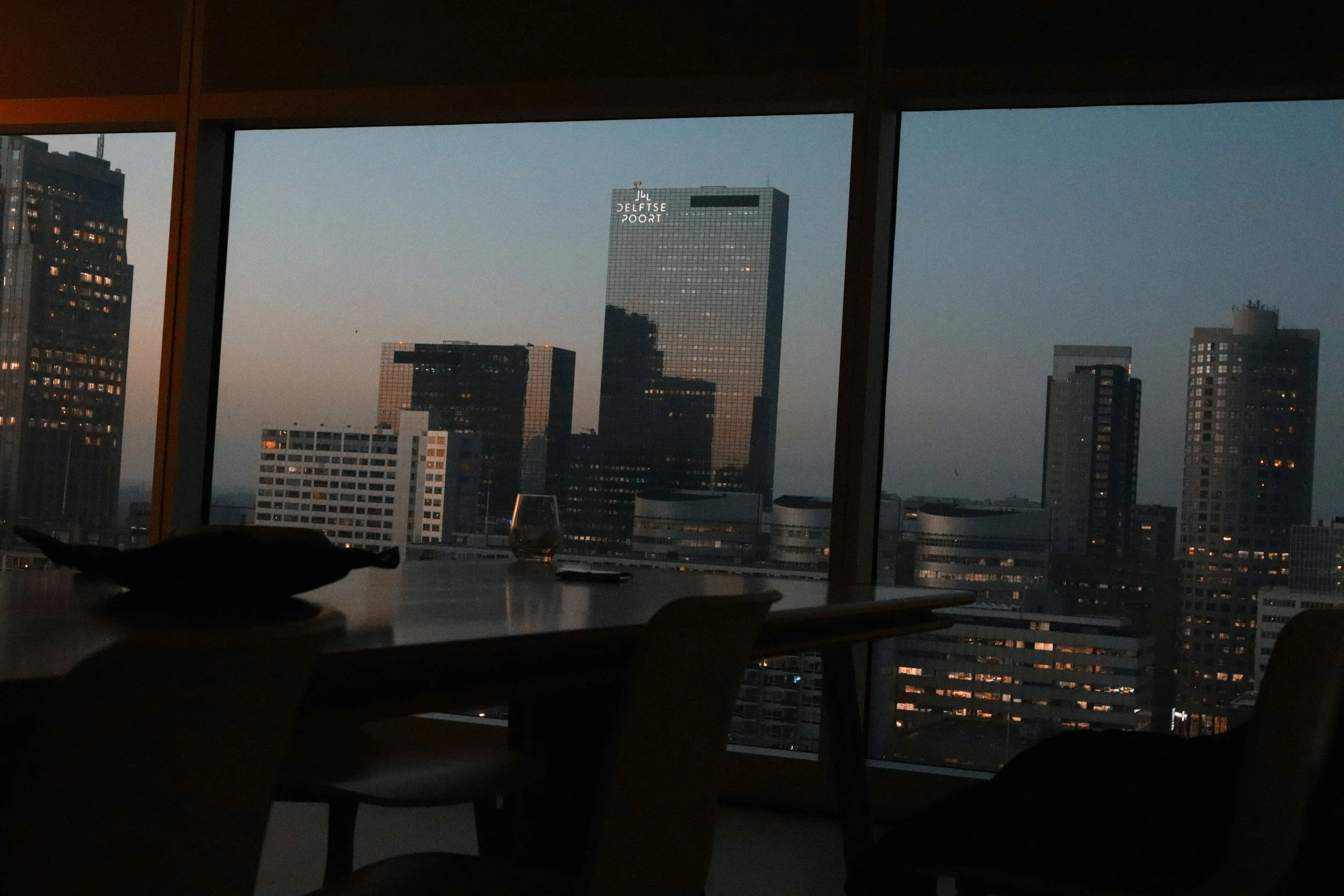 City skyline viewed from indoors at dusk, with a table and chairs in the foreground, and tall office buildings including one labeled 'DELFTSE POOIRT' in the background.