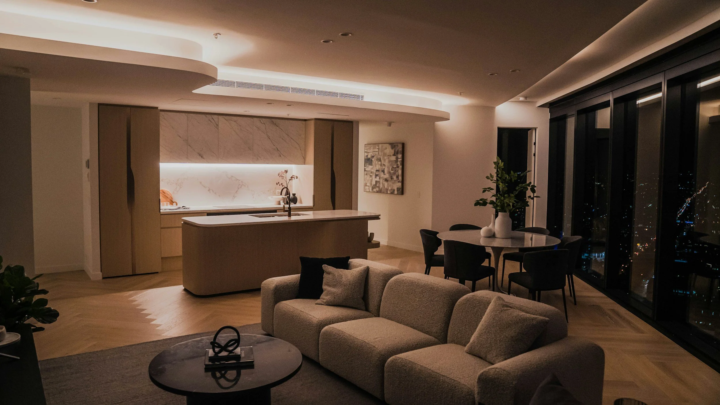 Living room and kitchen area in a modern apartment at night with city lights visible through large windows.
