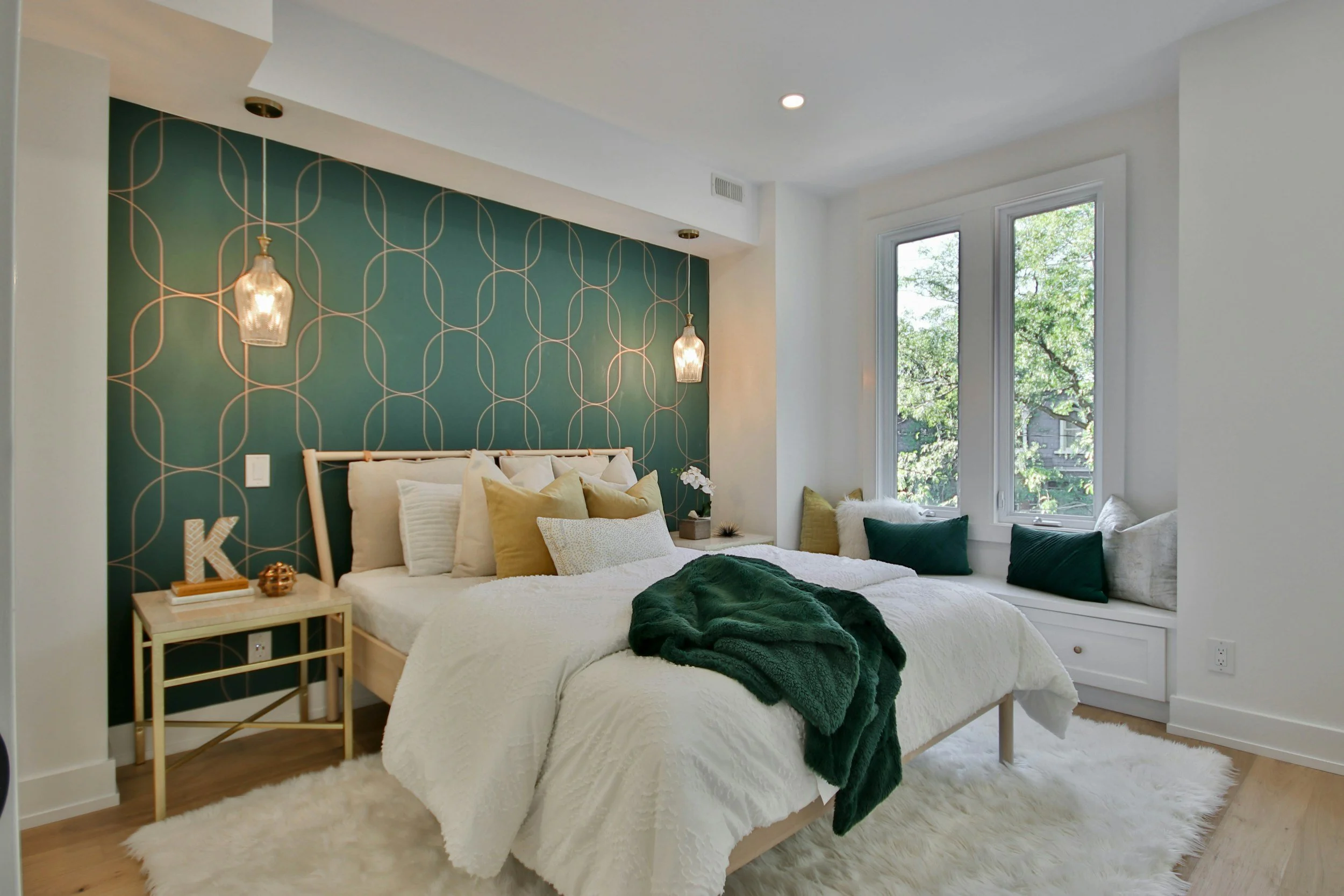 Cozy bedroom with a bed covered in white bedding and pillows, a dark green throw blanket, a small beige side table with decorative items, green and beige pillows on the window seat, and a teal accent wall with gold geometric pattern, pendant lights, 