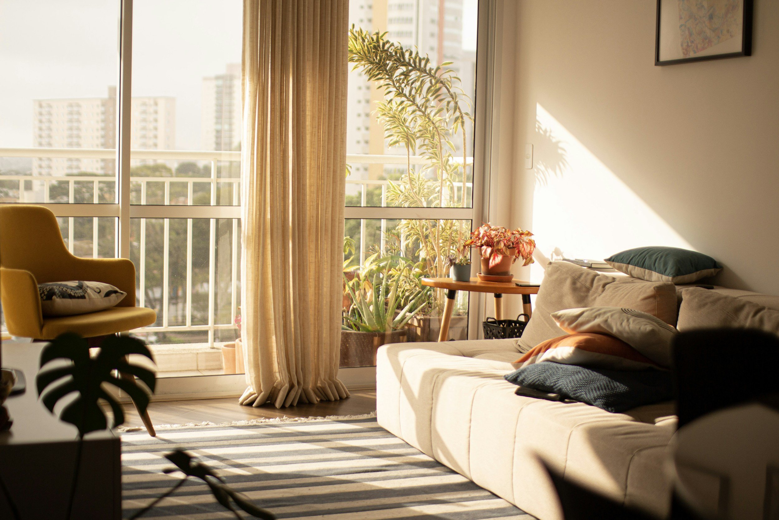 Bright living room with beige sofa, green pillow, and a side table with plants, large glass sliding doors with curtains, cityscape outside, and sunlight casting shadows.