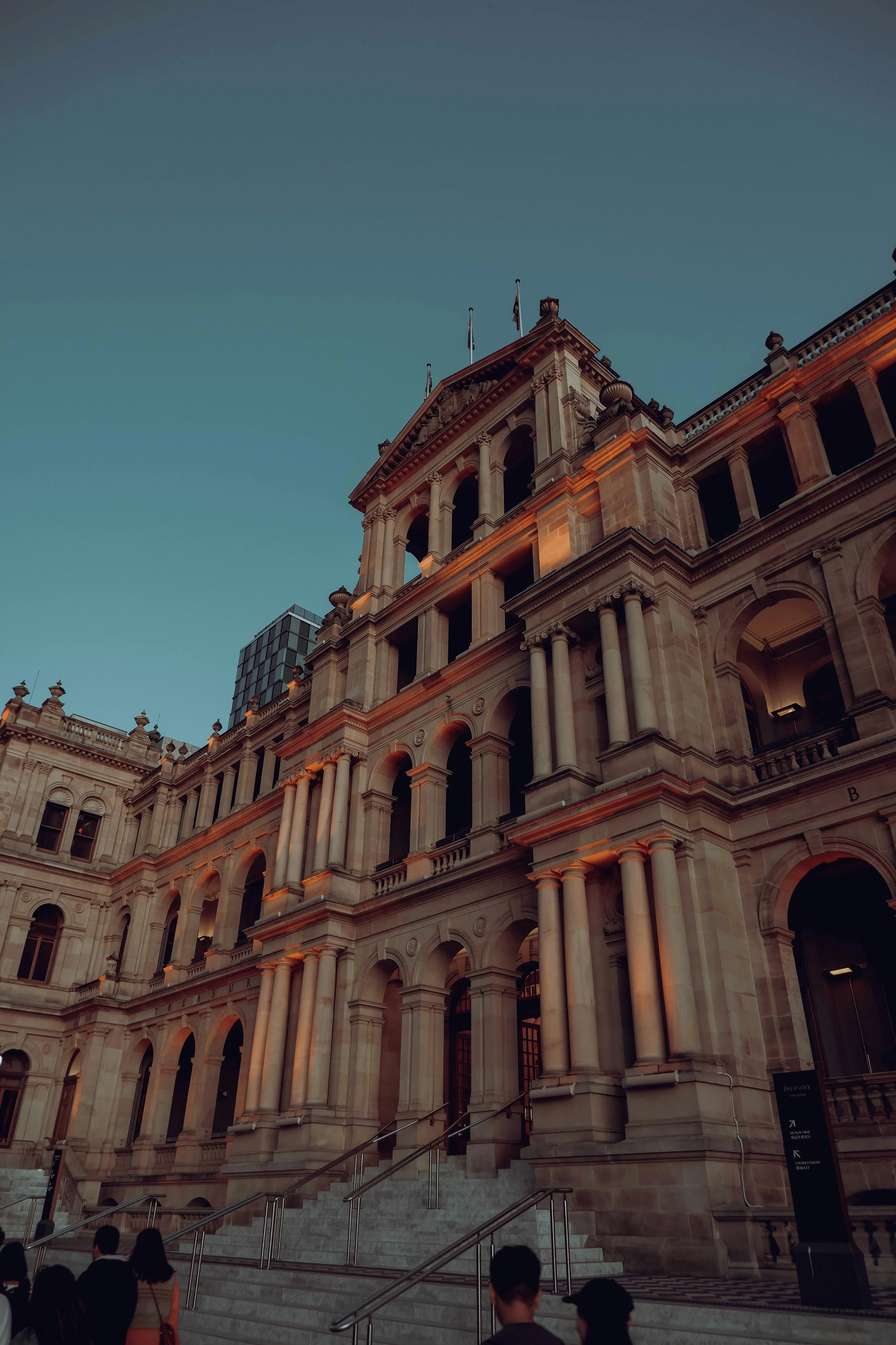 A historic building with classical architecture, stone columns, and arched windows, illuminated by warm lighting at sunset, with a few people standing at the bottom of stairs.