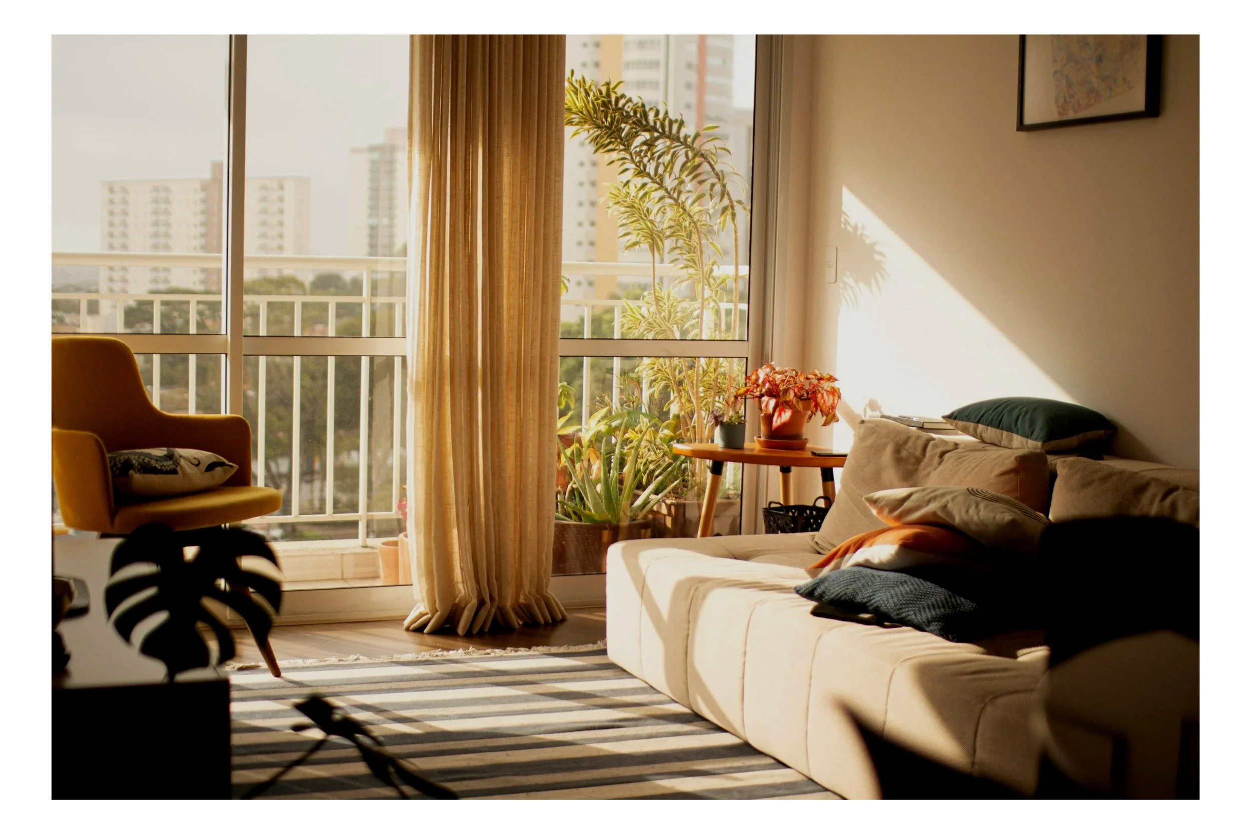 Sunlit living room with a beige sofa, multiple cushions, potted plants on a side table, large window with city views, yellow armchair, striped rug, and sunlight casting shadows inside.
