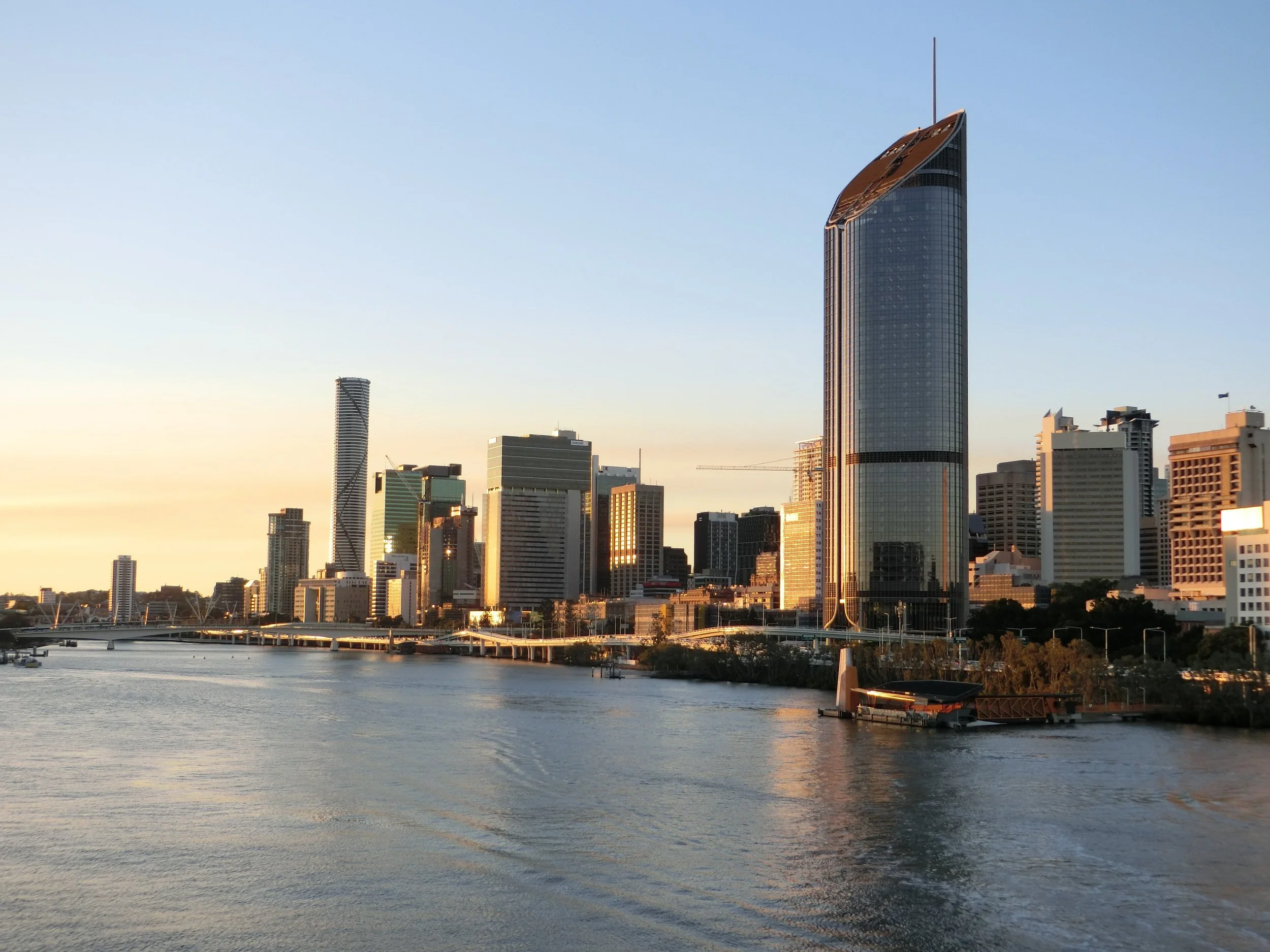 Skyline of a modern city with tall skyscrapers along a river at sunset, including a prominent glass tower with a curved top.