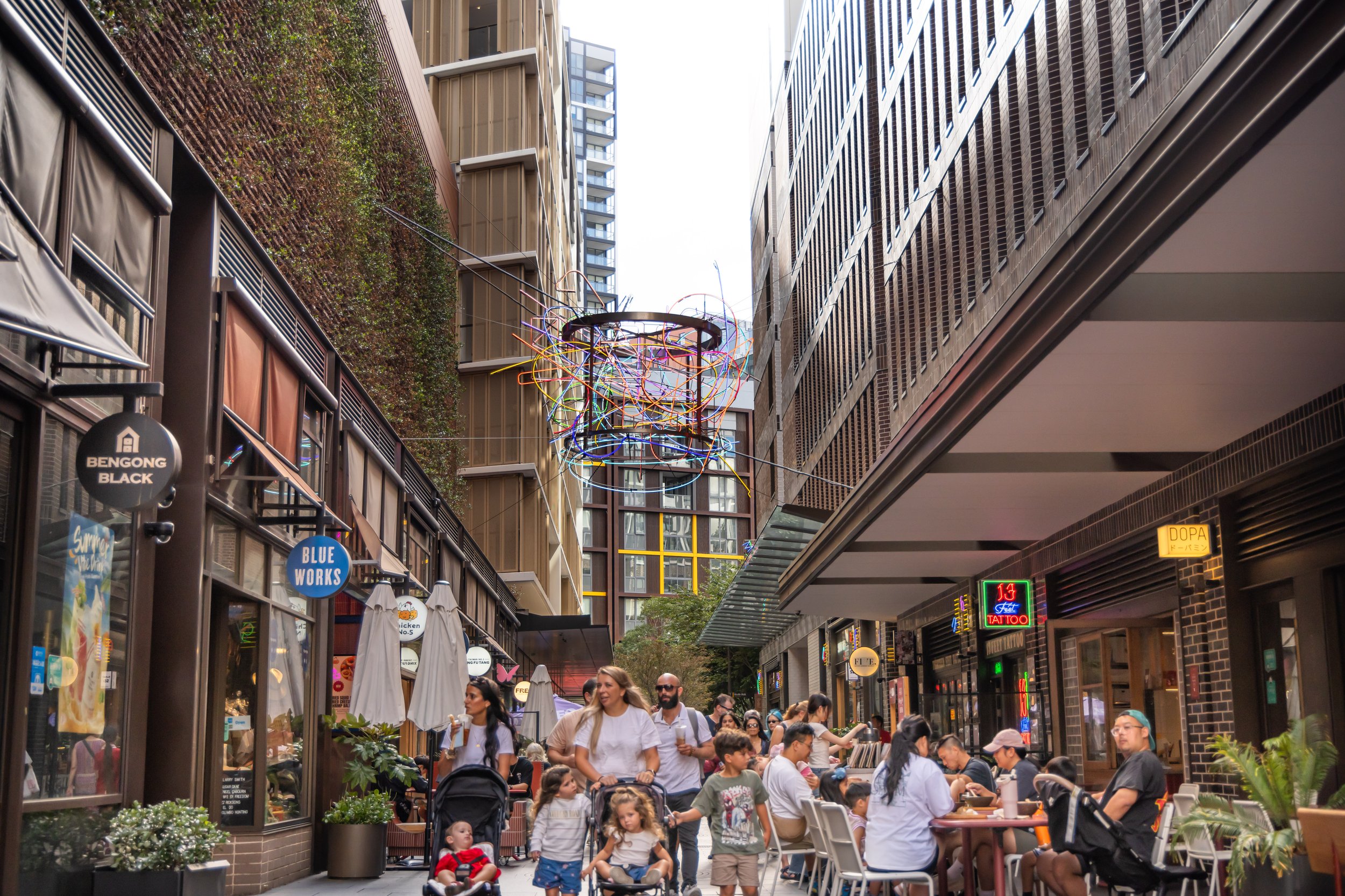 Urban street scene with people dining outside at cafes and walking along the sidewalk, surrounded by tall buildings and decorative street lighting.