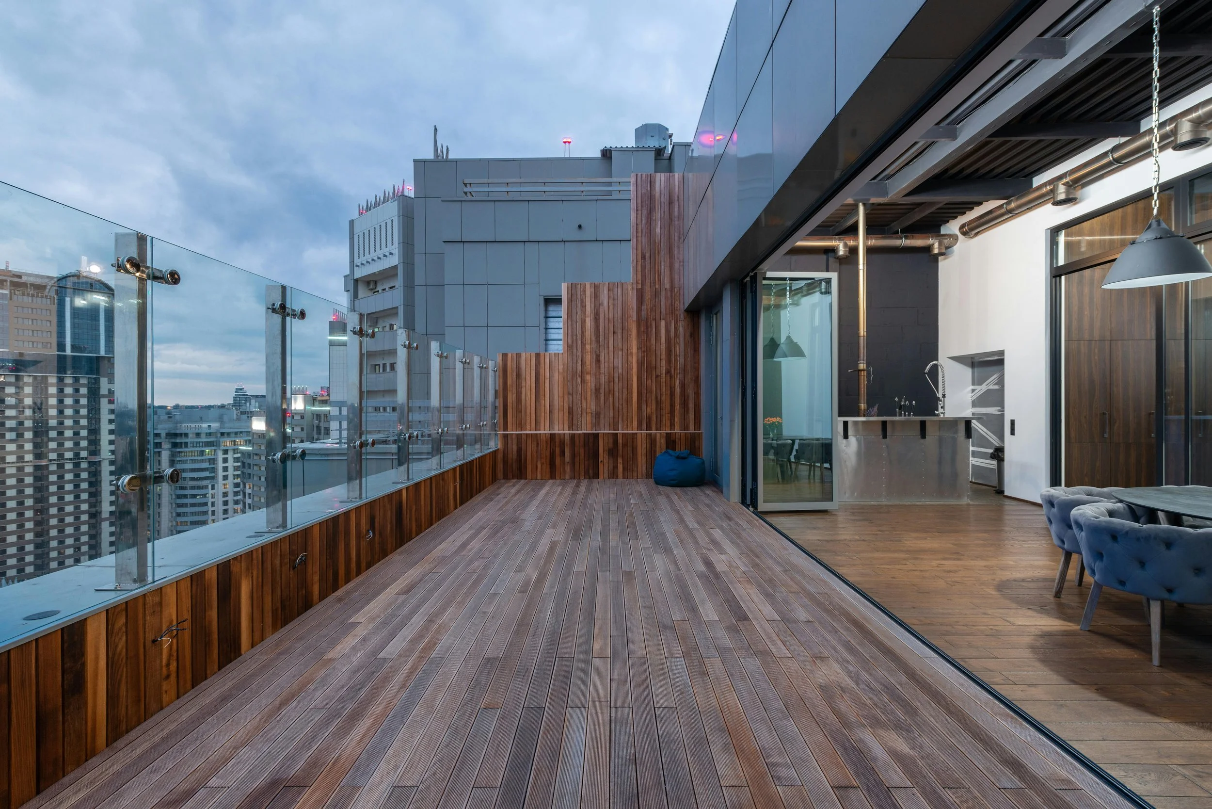 Urban rooftop terrace with wooden flooring, glass railing, city skyline, and an interior space visible through sliding glass doors, featuring a kitchen and dining area with tufted chairs.
