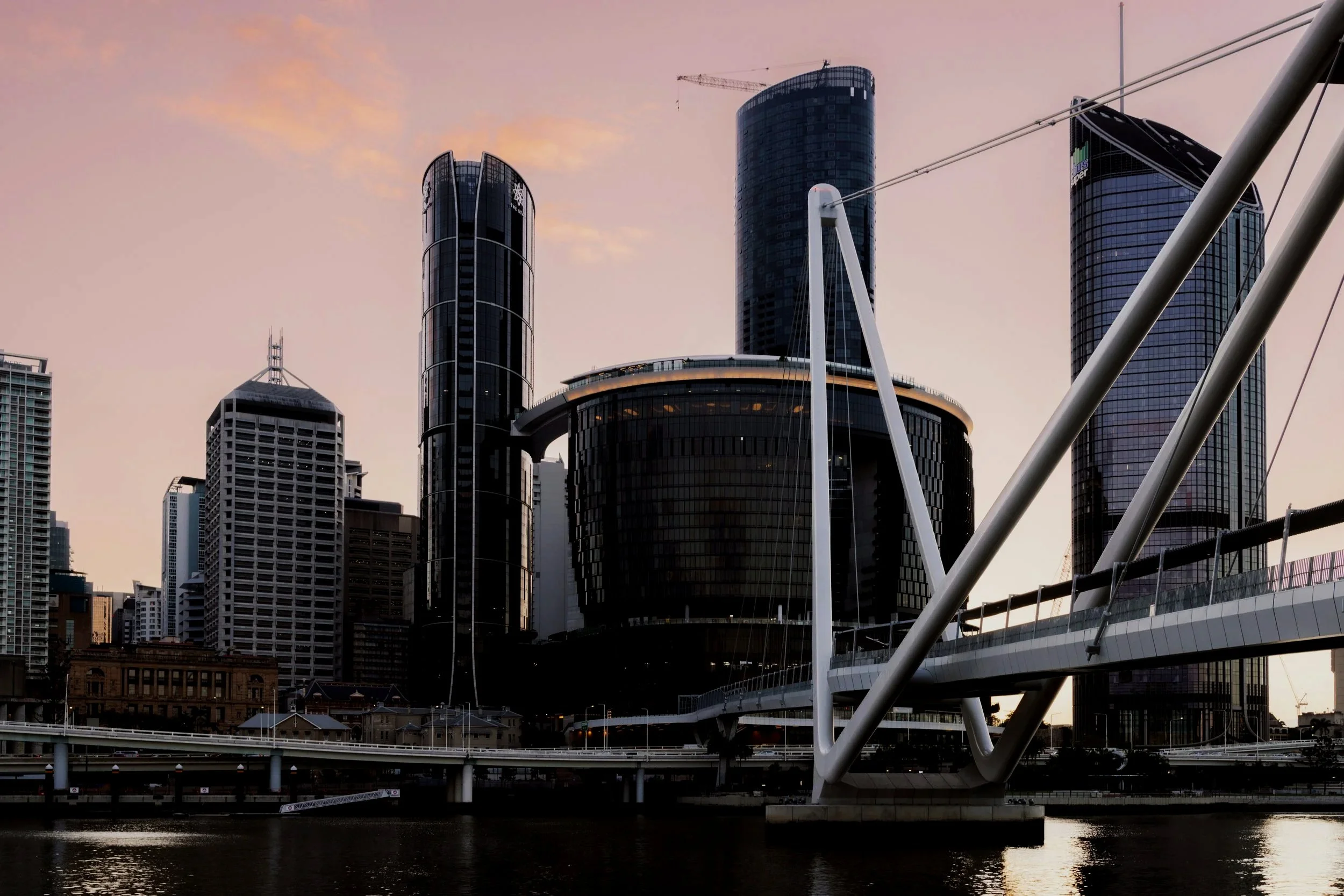 City skyline with modern high-rise buildings, bridge, and a body of water at sunset.