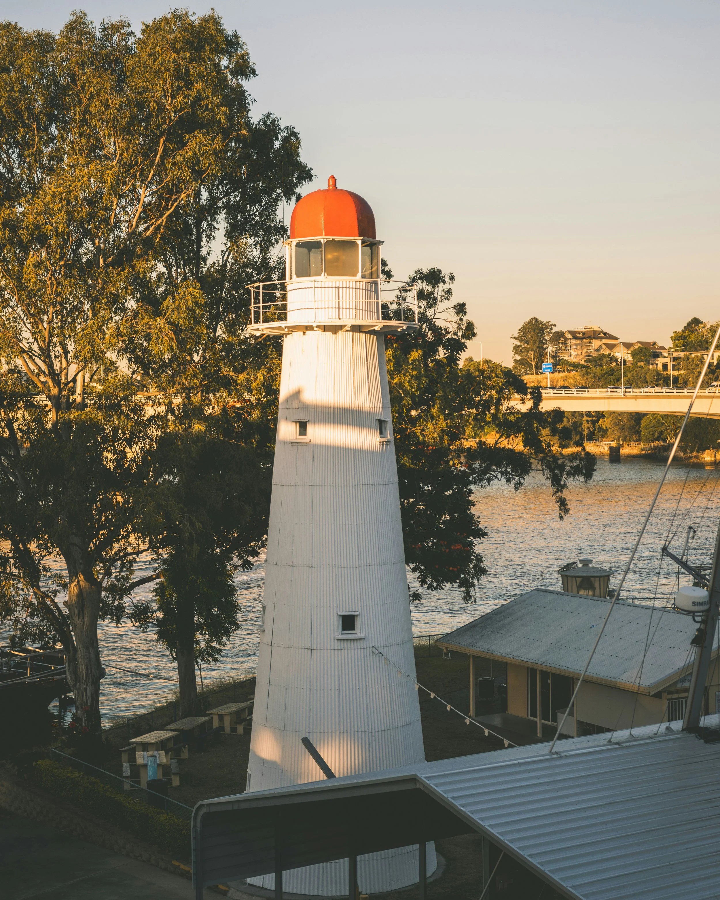 A white lighthouse with a red top next to a river, surrounded by trees and buildings in the background, during sunset.