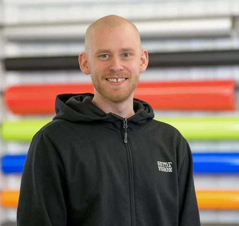 Ung man med ljus hud, kort blond hår, blont skägg och svart hoodie står framför färgade kanoter i ett lagerrum.