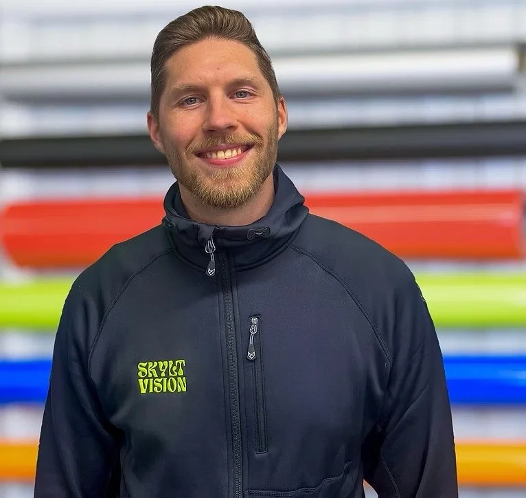 Ung man med ljus hy, ljusbrunt hår och skägg, ler, bär en svart jacka med texten 'Skylt Vision' och står framför en hylla med färgglada kajaker.