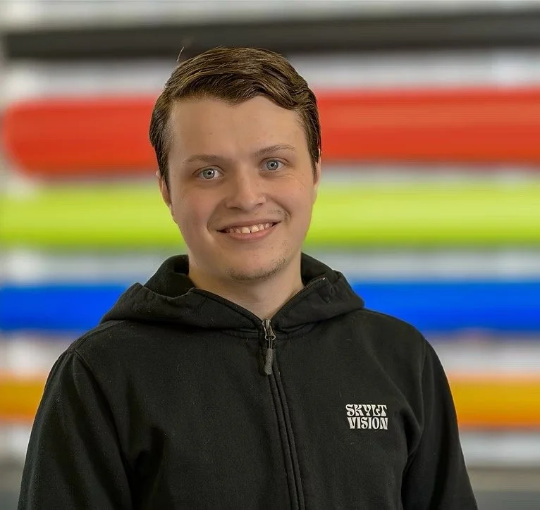 Ung man med ljus hy, kort brunt hår, och blå ögon, som bär en svart hoodie med texten 'SKYDER VISION', står framför ett bakgrund med färgglada band.