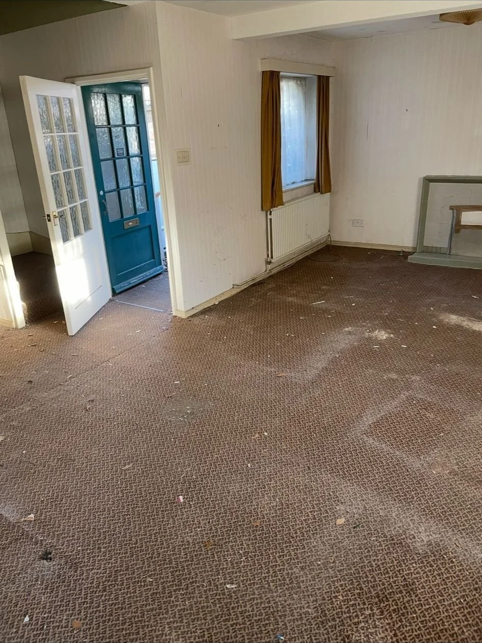 Empty room with brown carpet, white walls, a window with yellow curtains, and a blue front door open to the outside.